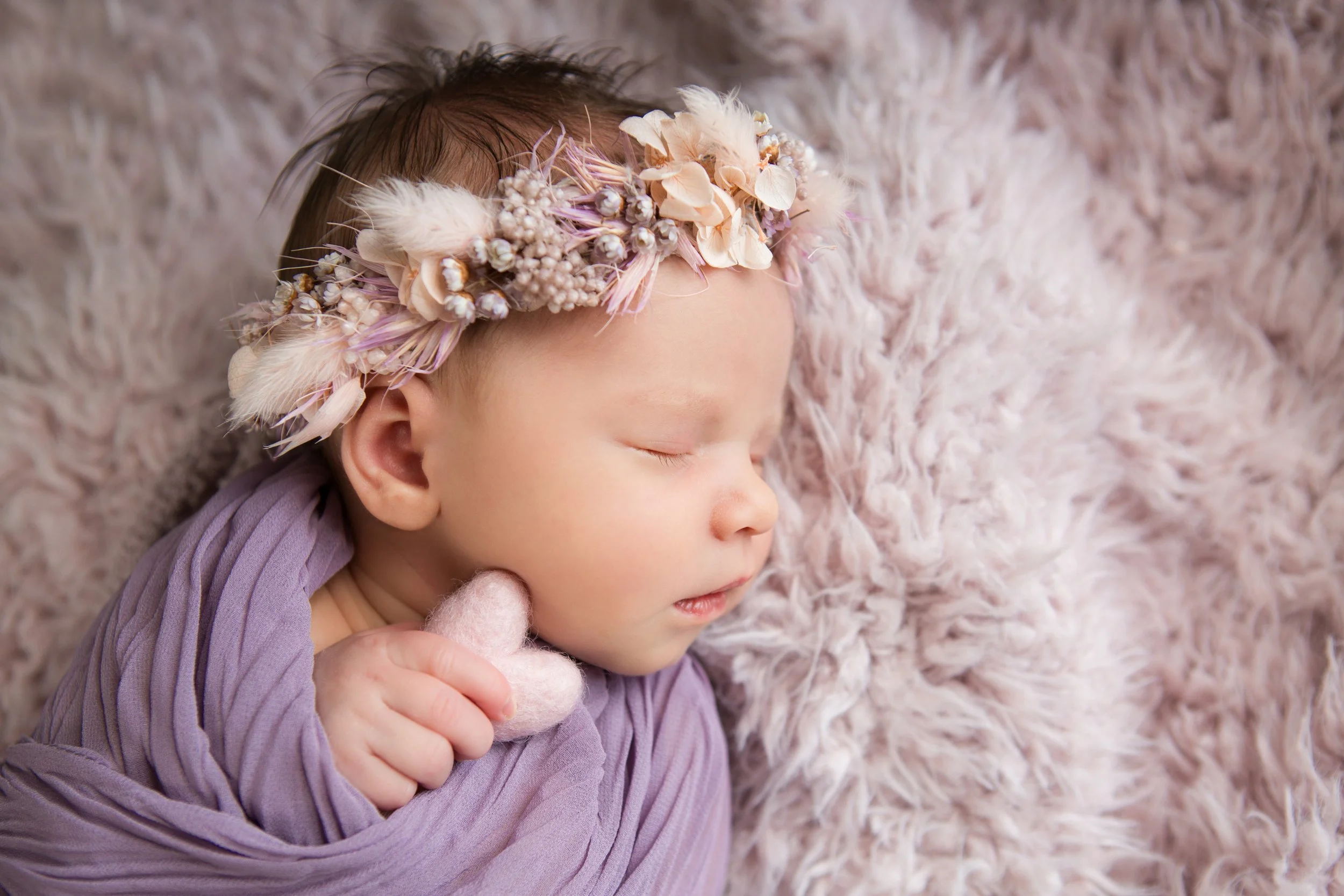 Baby Girl with a flower and feather headband holding a plush heart wrapped in purple
