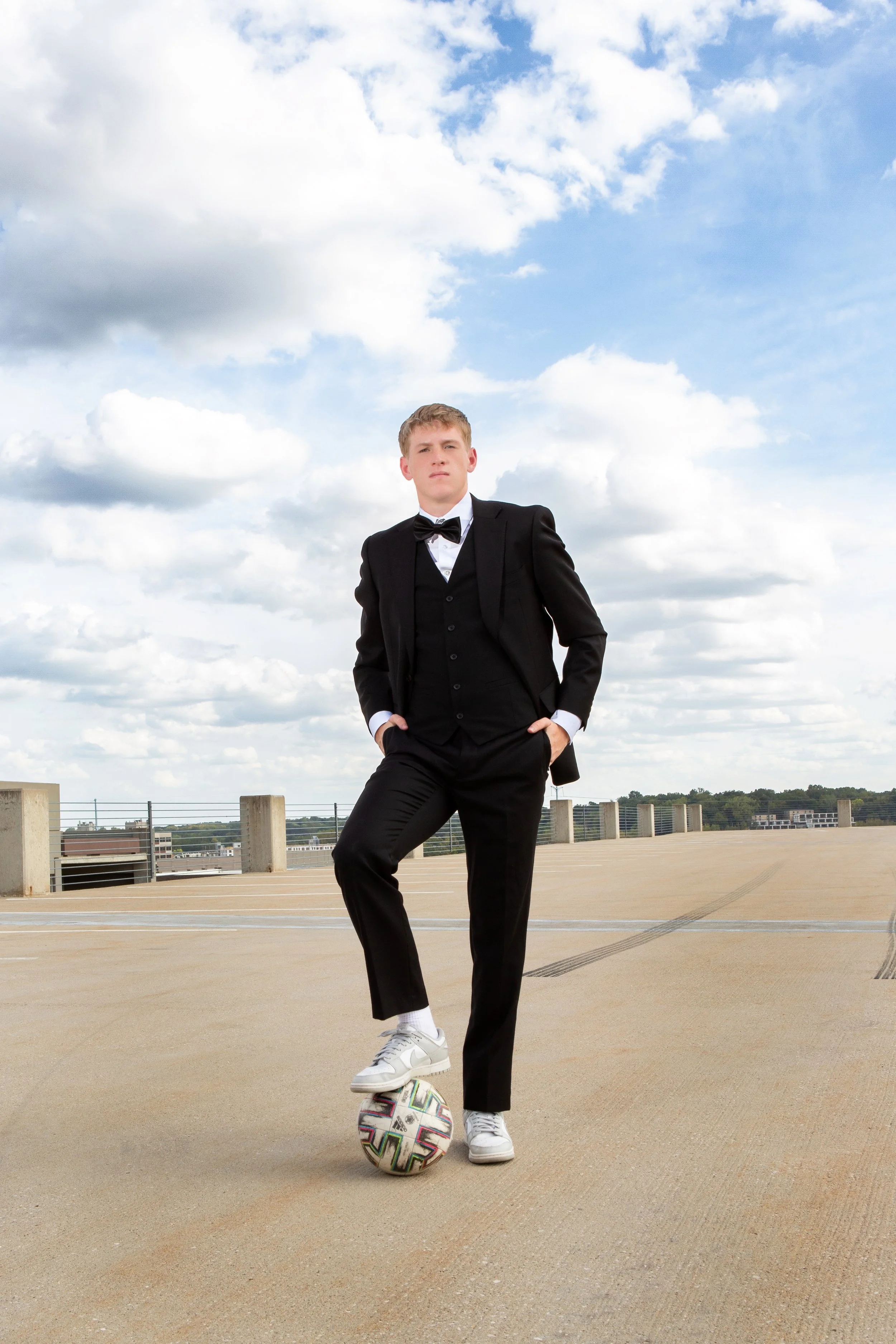 A young man in a black tuxedo with a bow tie stands confidently on a rooftop parking lot, with one foot on a colorful soccer ball, against a backdrop of a cloudy blue sky.
