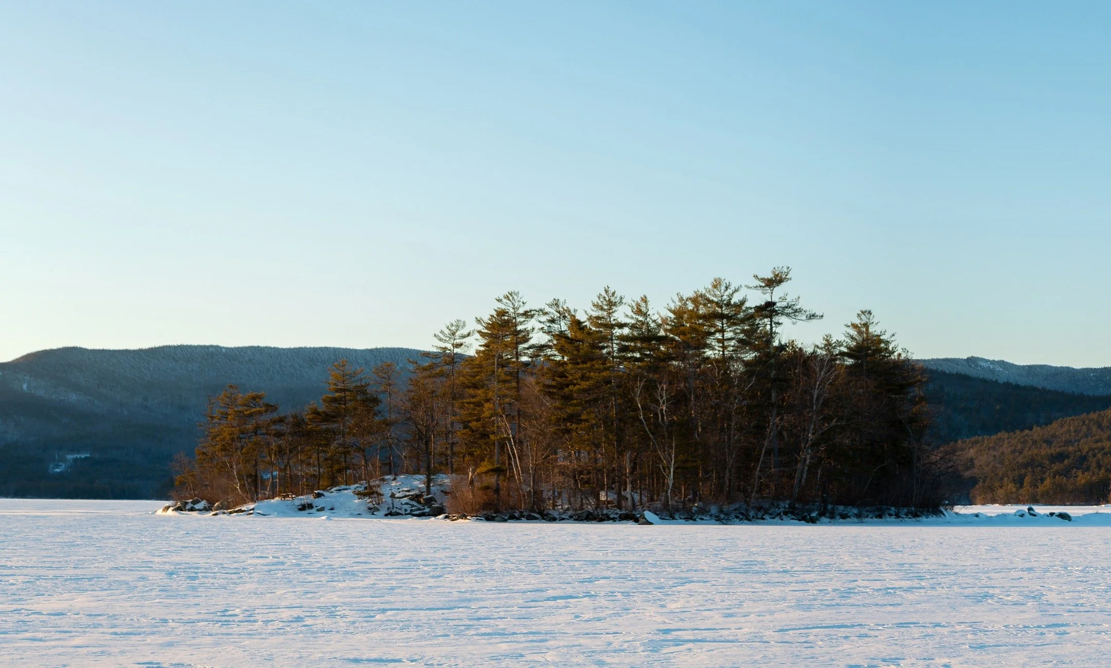 Loon Island casting a long evening shadow across a frozen Squam Lake. In the distance you can see East Rattlesnake Mountain and behind that the Squam Range.