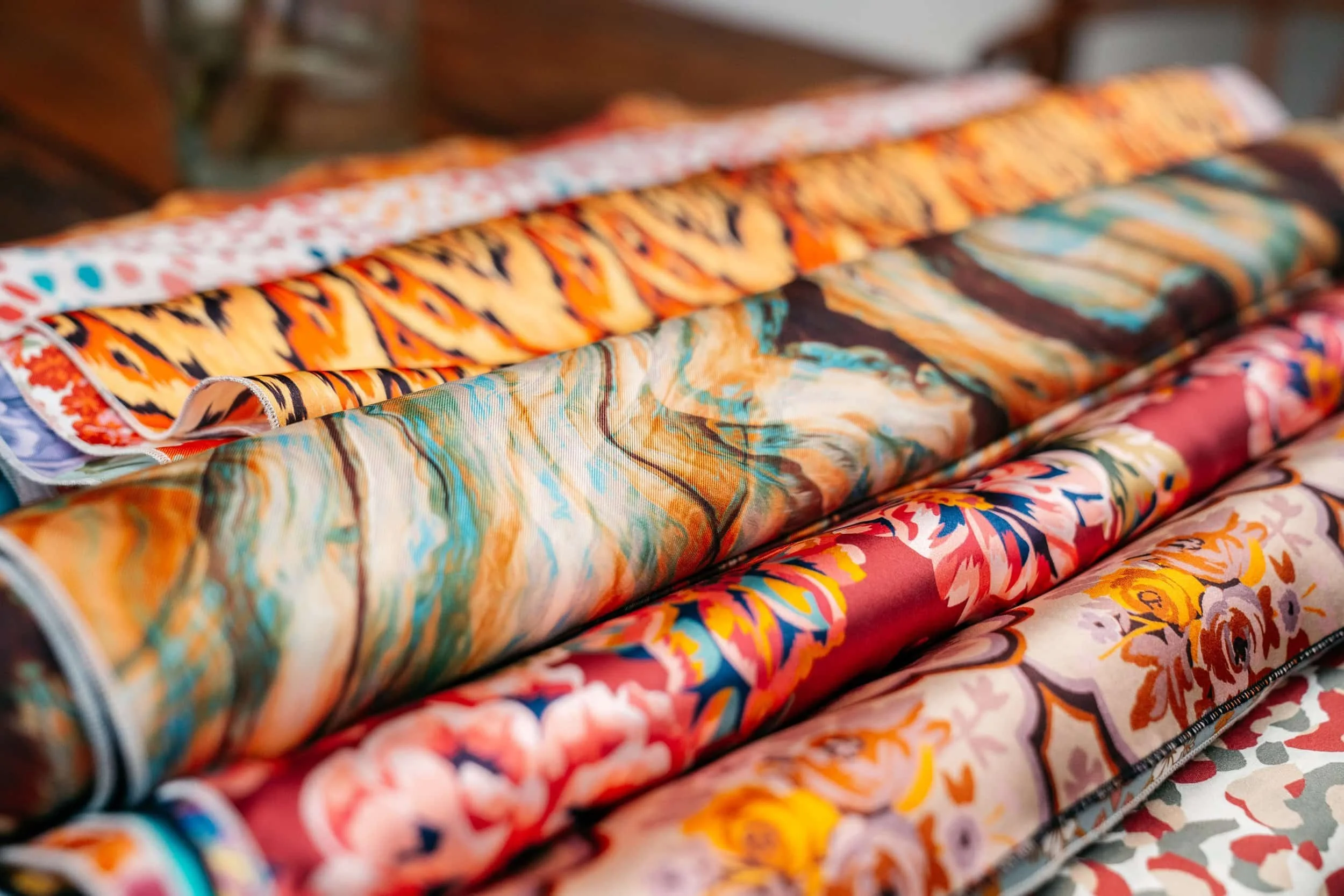 Rolls of colorful patterned fabric with floral, abstract, and tiger-stripe designs stacked on a wooden surface.