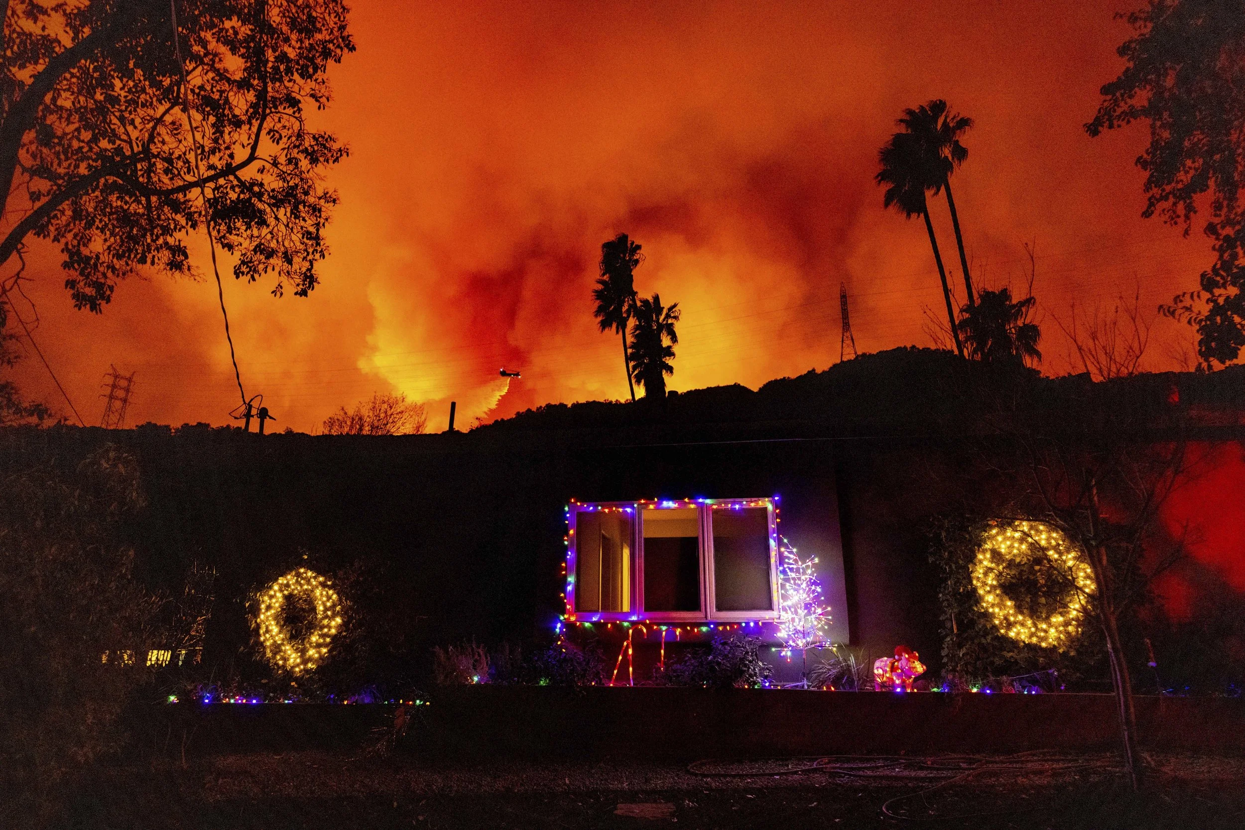  A helicopter drops water on the Palisades Fire behind a home with Christmas lights in Mandeville Canyon, Friday, Jan. 10, 2025, in Los Angeles. (AP Photo/Ethan Swope) 