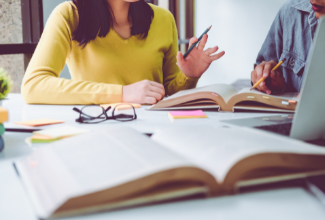 A woman with a yellow shirt, talking with another person in front of several books and a computer