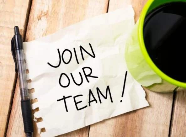 Join our team written on a paper next to a pen and a bright green coffee mug