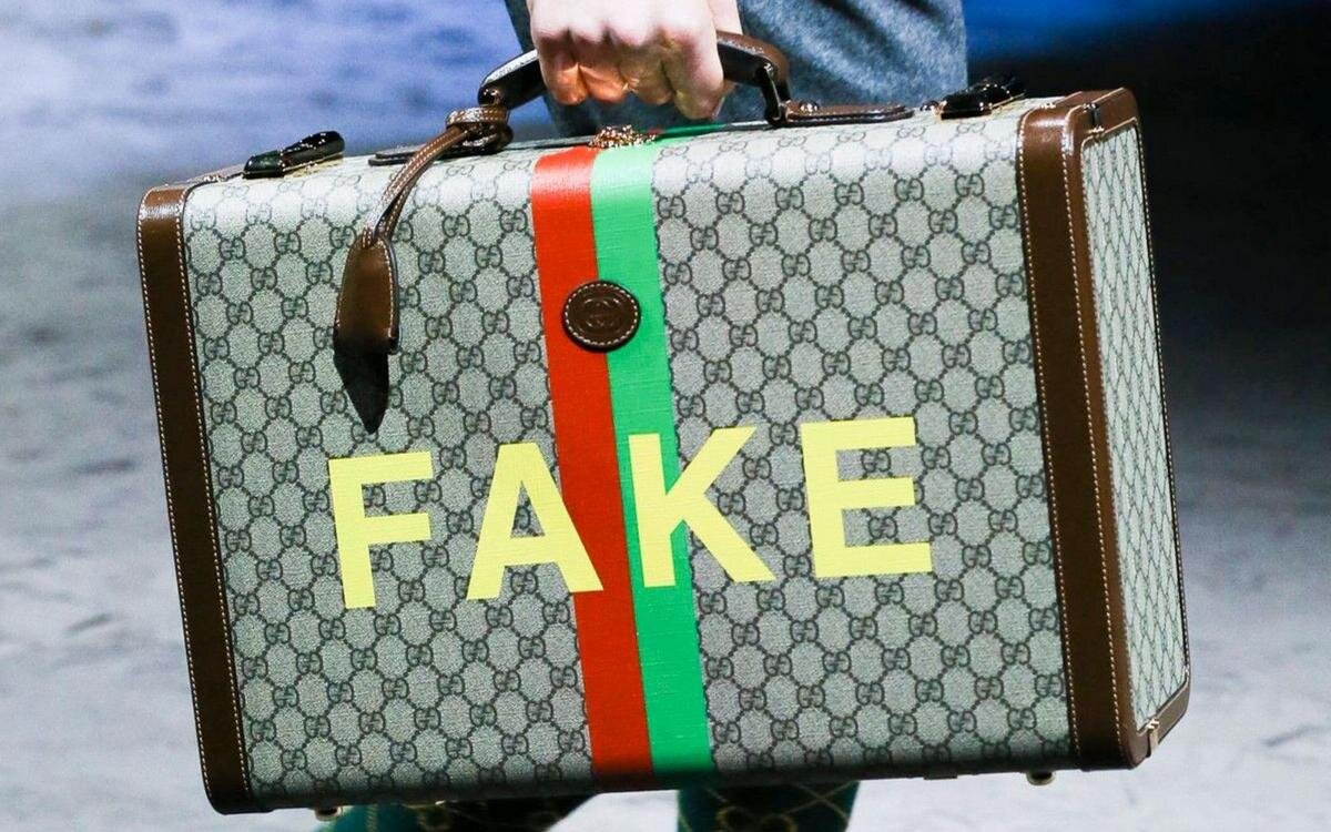 A hard suitcase from the Fake/Not capsule collection, first unveiled in Gucci’s AW20 show, prominently displaying ‘FAKE’ against Gucci’s iconic bicolor striped motif. Image Via
