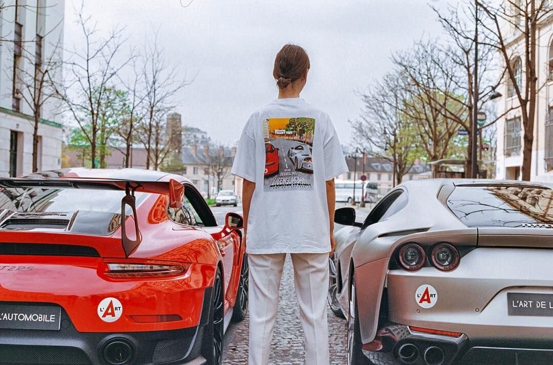 A model wears a L’ART tee depicting the exact scene she is standing in, featuring a Porsche 911 GT2RS and Ferrari 812 Superfast. Image  Via