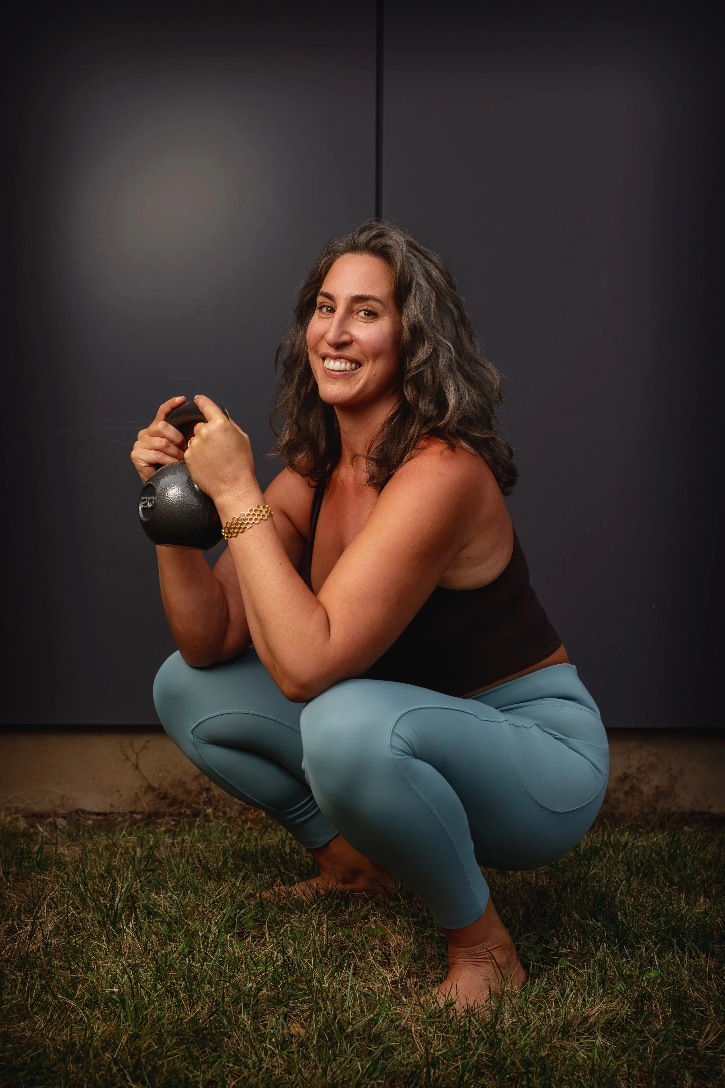 Smiling woman in workout clothes holding a kettlebell while exercising outdoors on grass