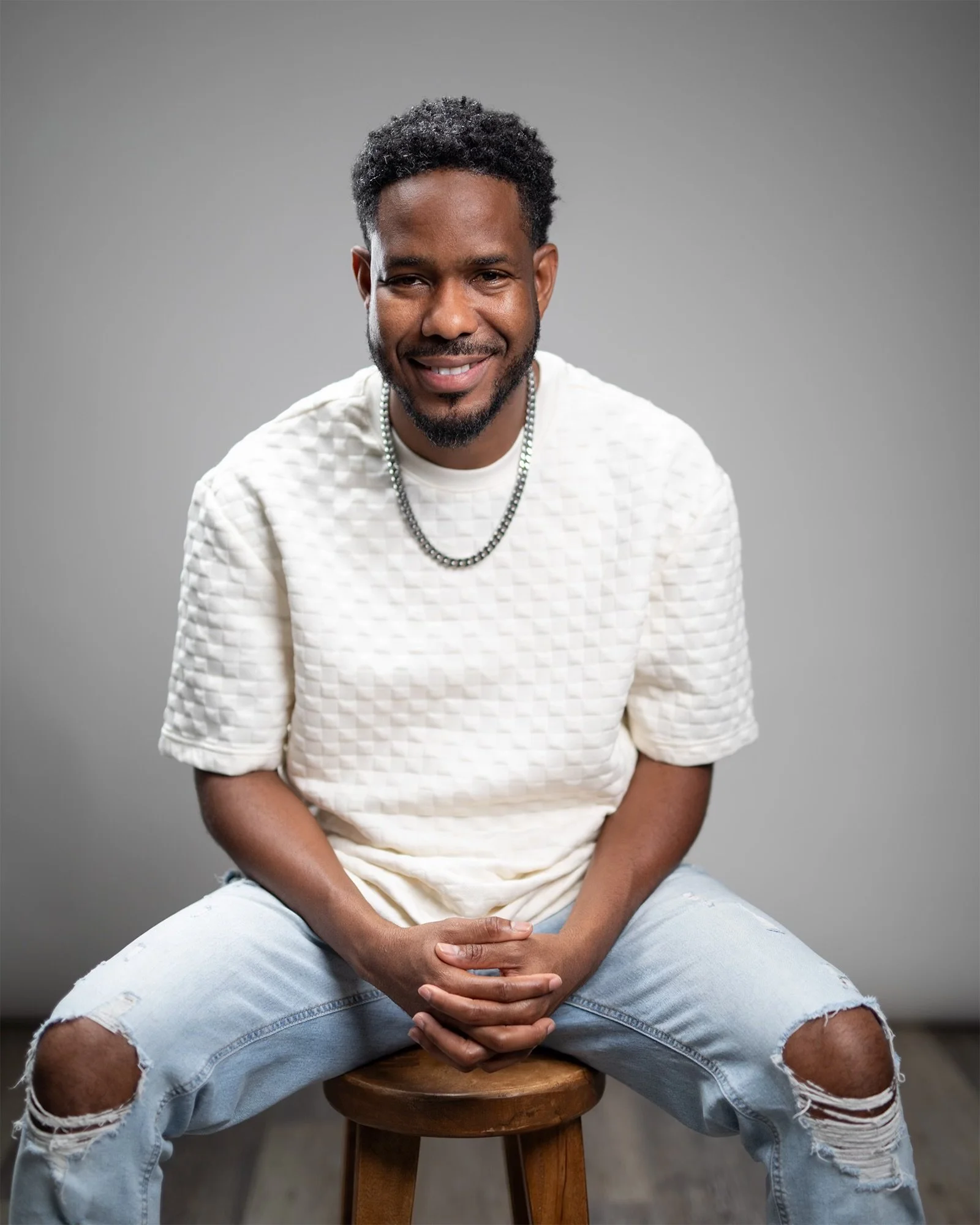 A man sitting on a stool, smiling, wearing a white textured shirt, light blue ripped jeans, a silver chain, and having curly black hair and a beard, against a plain gray background.
