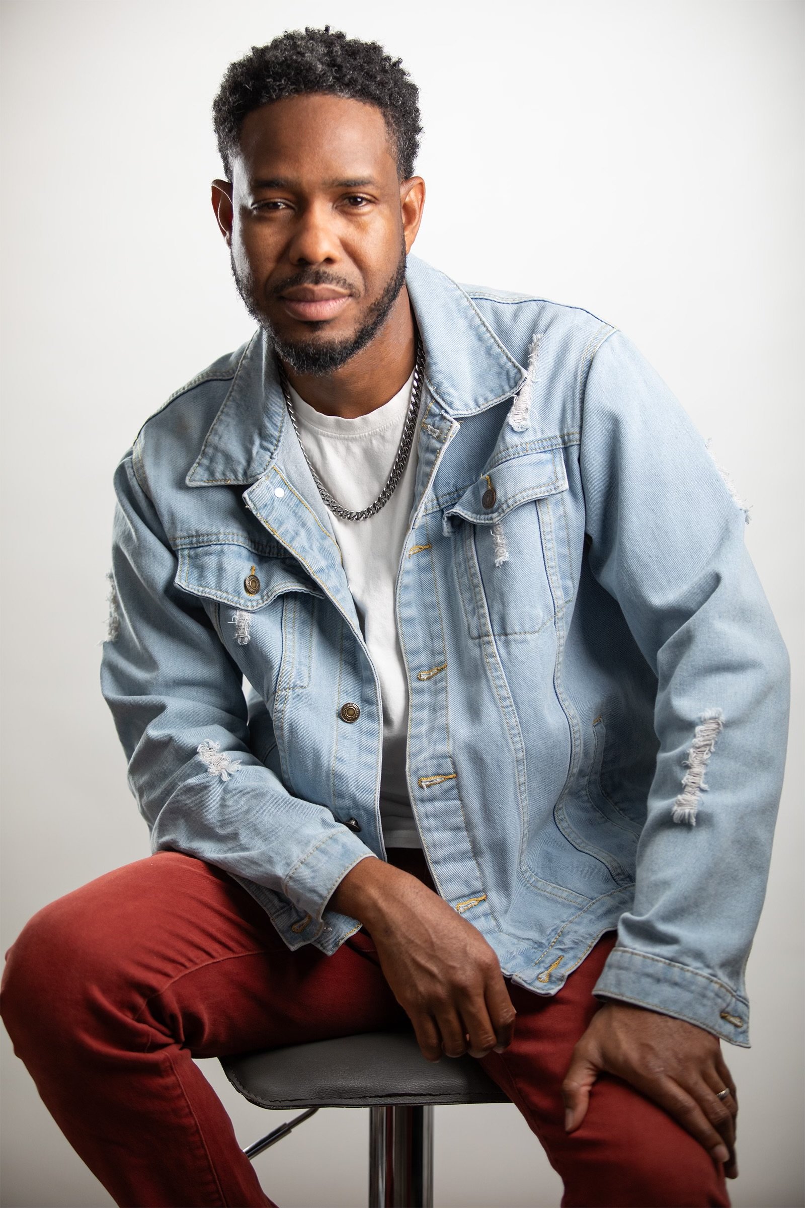 A man sitting on a stool against a white background, wearing a light blue denim jacket, white t-shirt, and red pants.