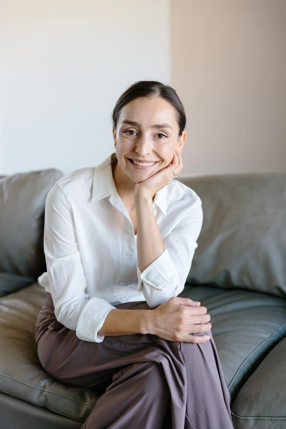 Jamie Mueller seated on a sofa in a white blouse and brown trousers, leaning forward and smiling.