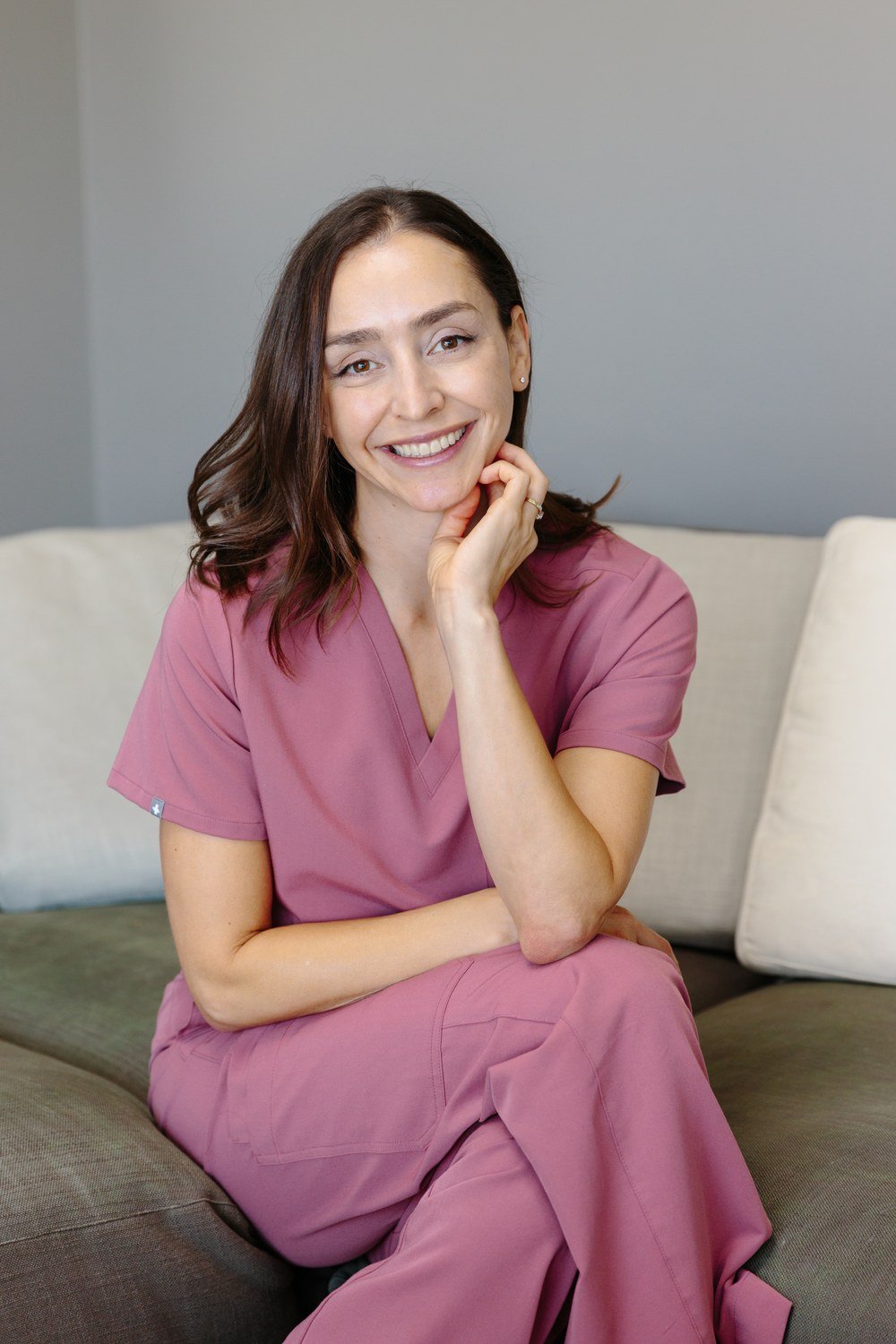 Jamie Mueller seated on a sofa in pink scrubs, smiling with her chin resting on her hand.