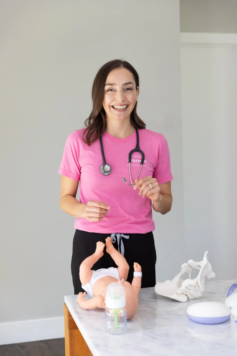 Jamie Mueller standing behind a table with baby feeding equipment, wearing a pink shirt and stethoscope.