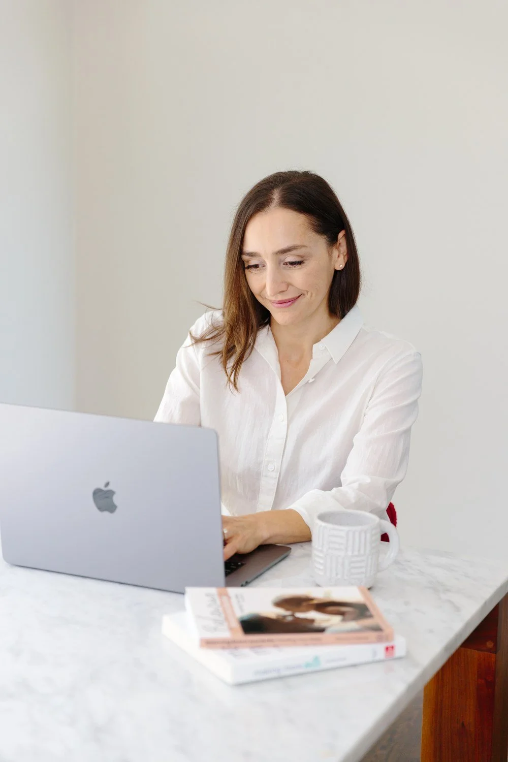 Jamie Mueller working at a laptop at a marble table, with a mug and books beside her.