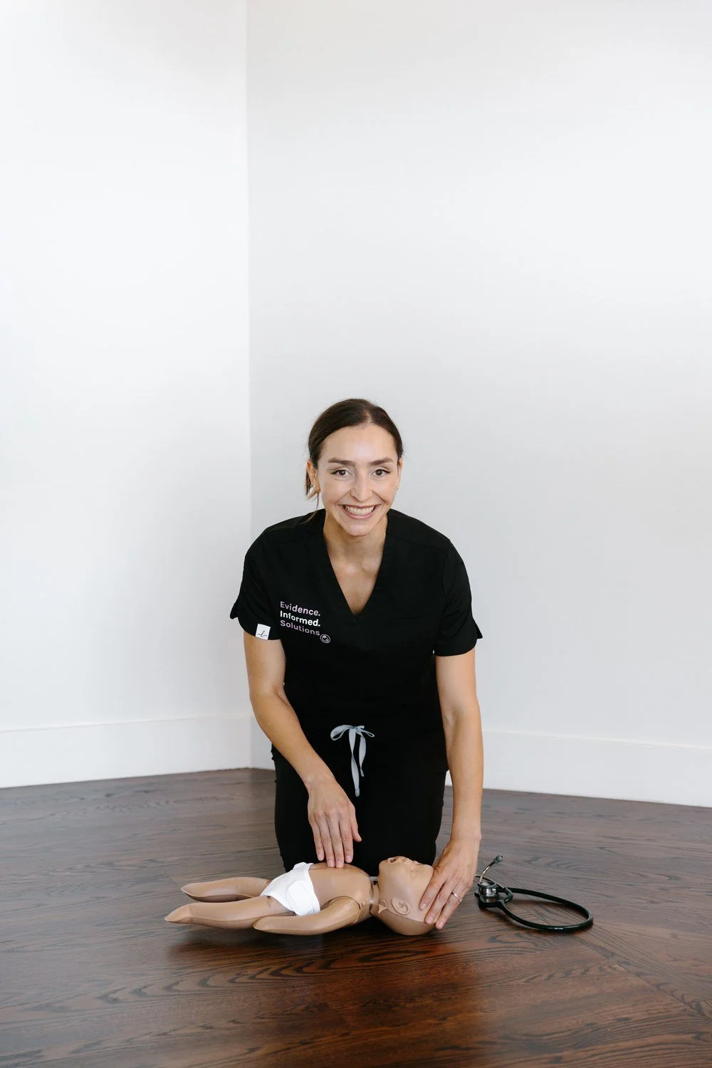 Jamie Mueller kneeling beside an infant mannequin on a wooden floor, with a stethoscope nearby.