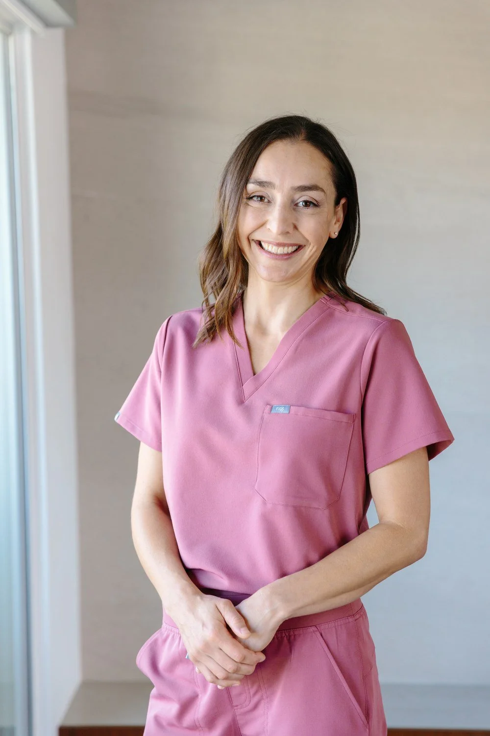 Jamie Mueller smiling beside a window in pink scrubs, with her hands resting in front of her.
