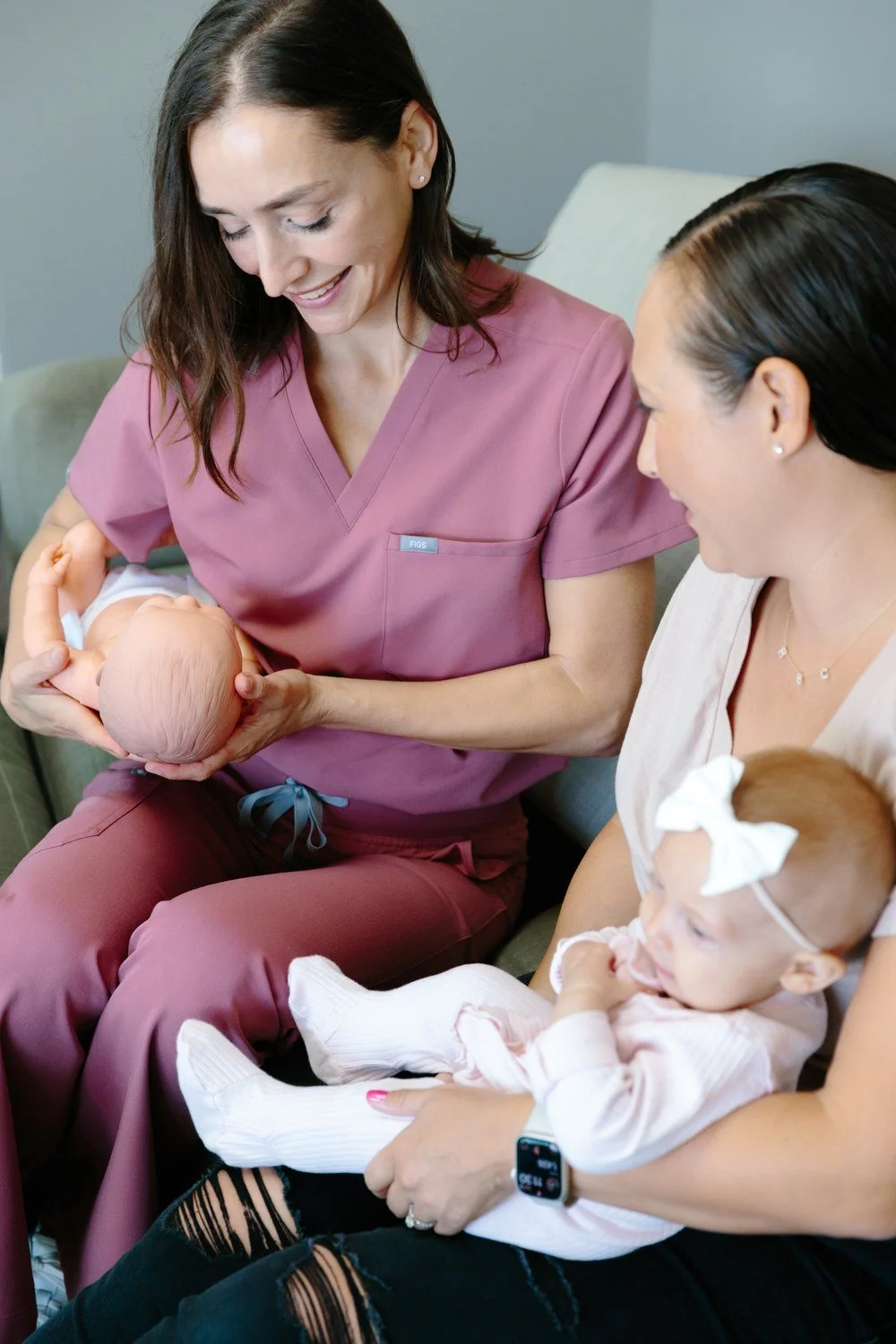 Jamie Mueller holding a baby doll while a seated woman holding an infant watches the demonstration.