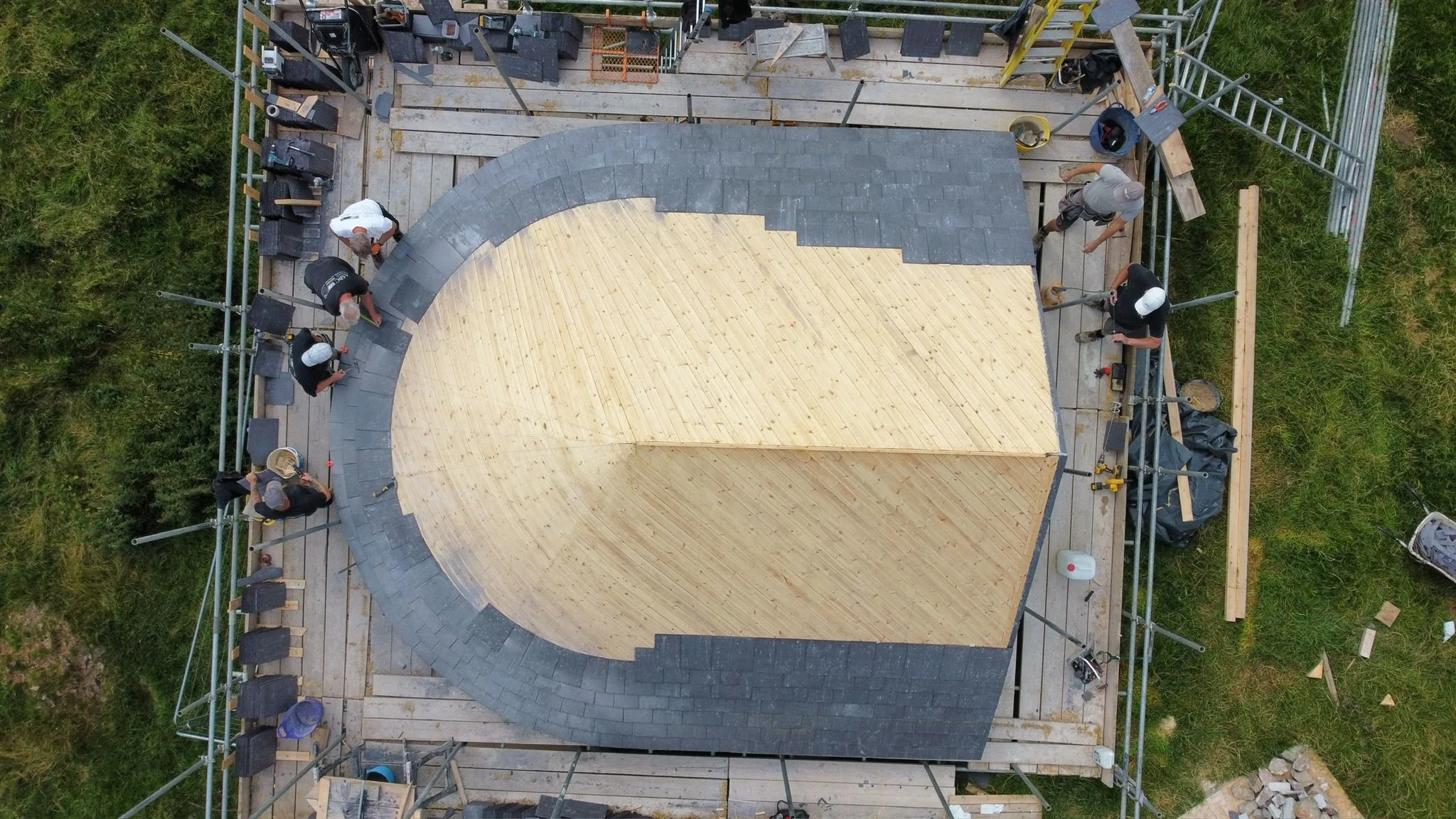 Aerial view of a construction site where workers are building a circular wooden structure, with parts of the roof covered with gray shingles, surrounded by scaffolding and green grass.
