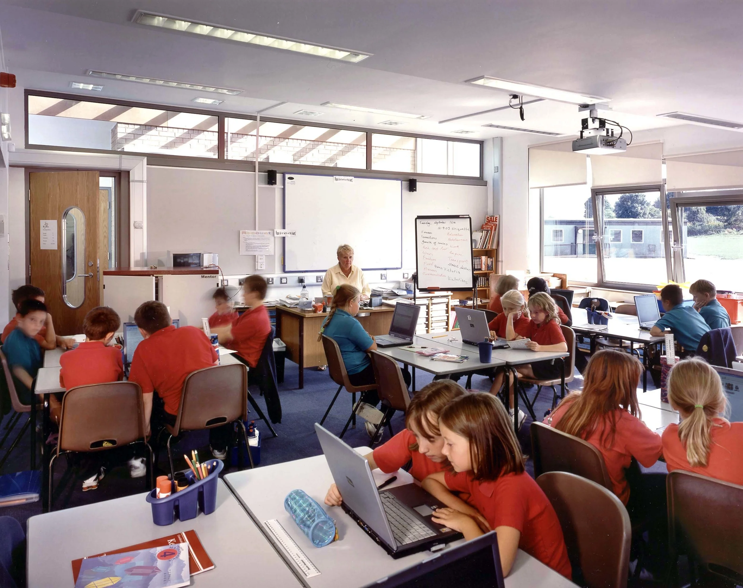 A classroom filled with students working on laptops at desks, with a teacher sitting in front of the class, whiteboard, windows, and various classroom supplies.