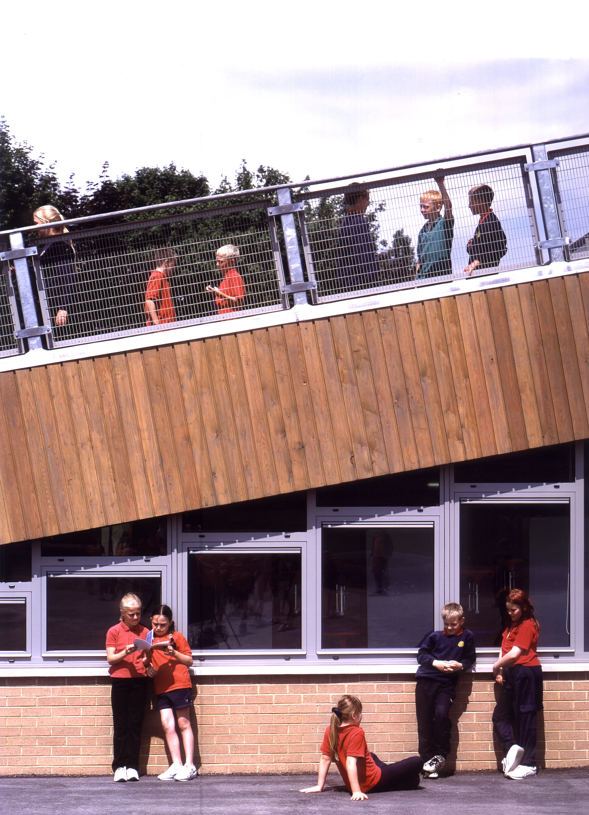 Group of children and teachers standing and sitting outside in front of a building, with some on the lower ground and some on a balcony, engaging in activities and conversations on a sunny day.