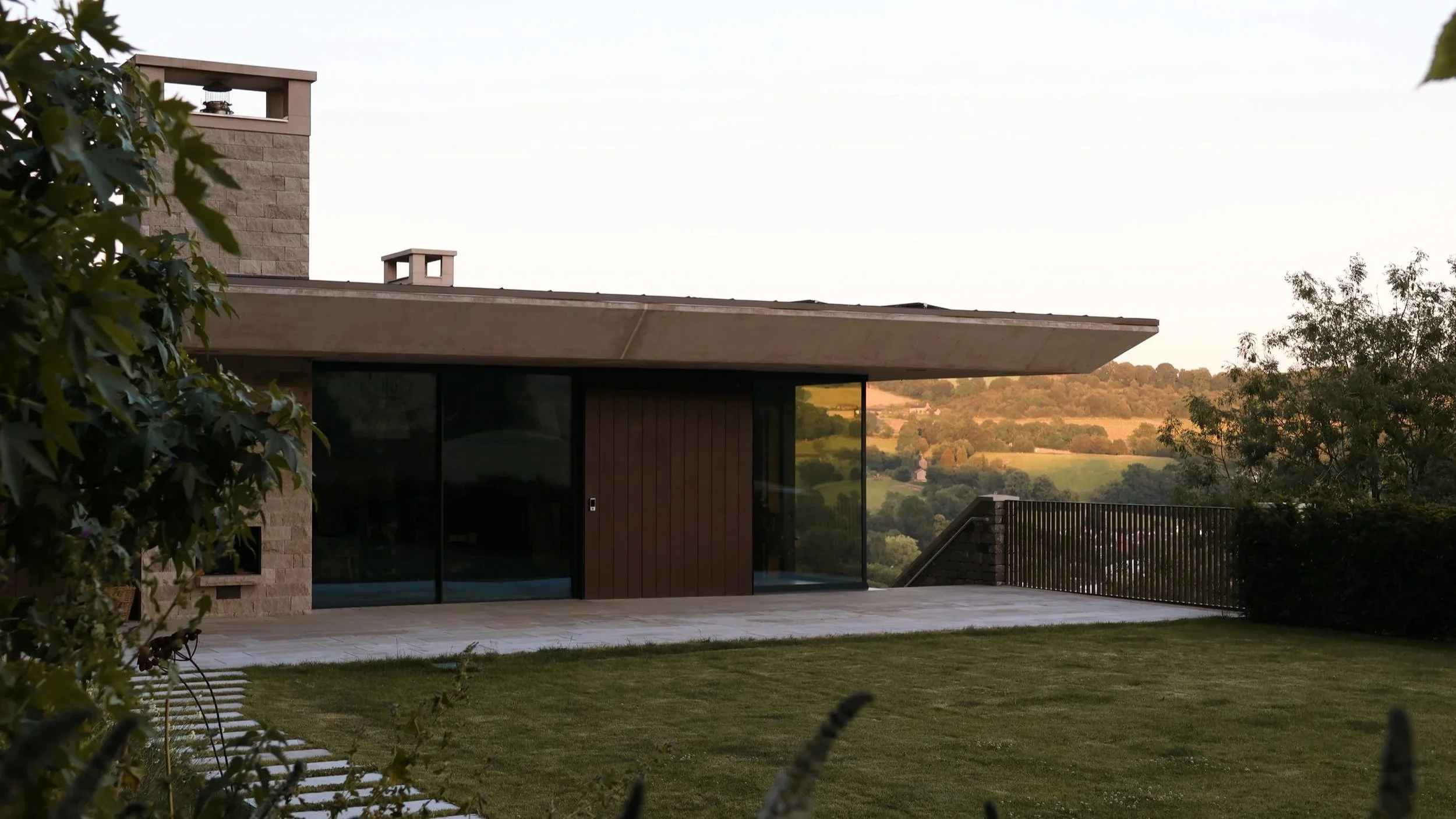 Modern house exterior with large sliding glass door, stone and wooden facade, adjacent patio, green lawn, trees, and hills in the background at sunset.