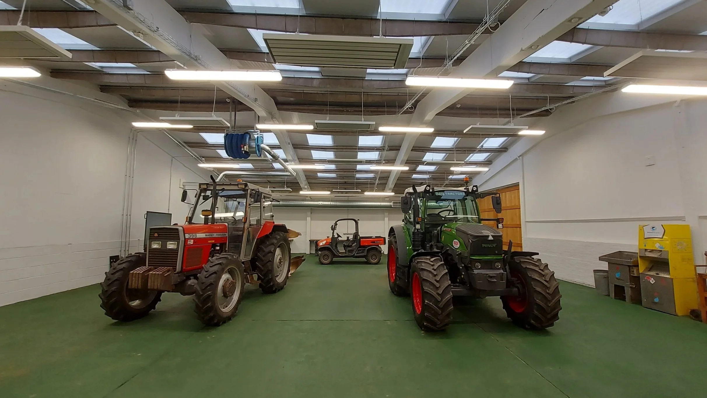 Indoor display of farm equipment, including red and green tractors and smaller utility vehicles, with a ceiling featuring skylights and fluorescent lighting.