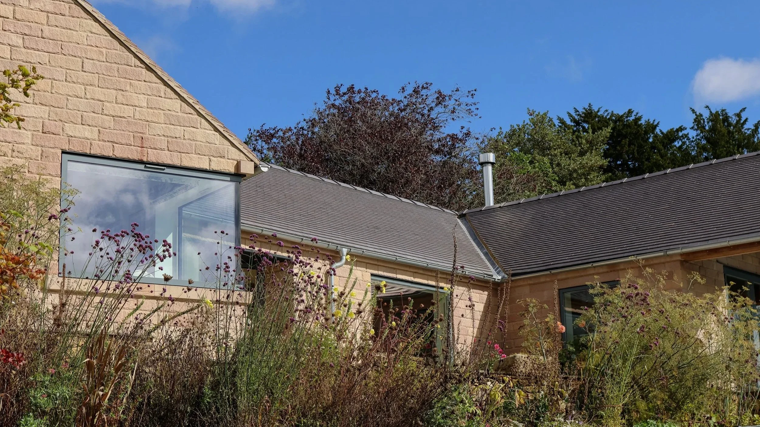 A house with beige brick walls and a dark gray shingle roof, with a large window and a garden with flowering plants in the foreground, set against a bright blue sky with some clouds and trees in the background.