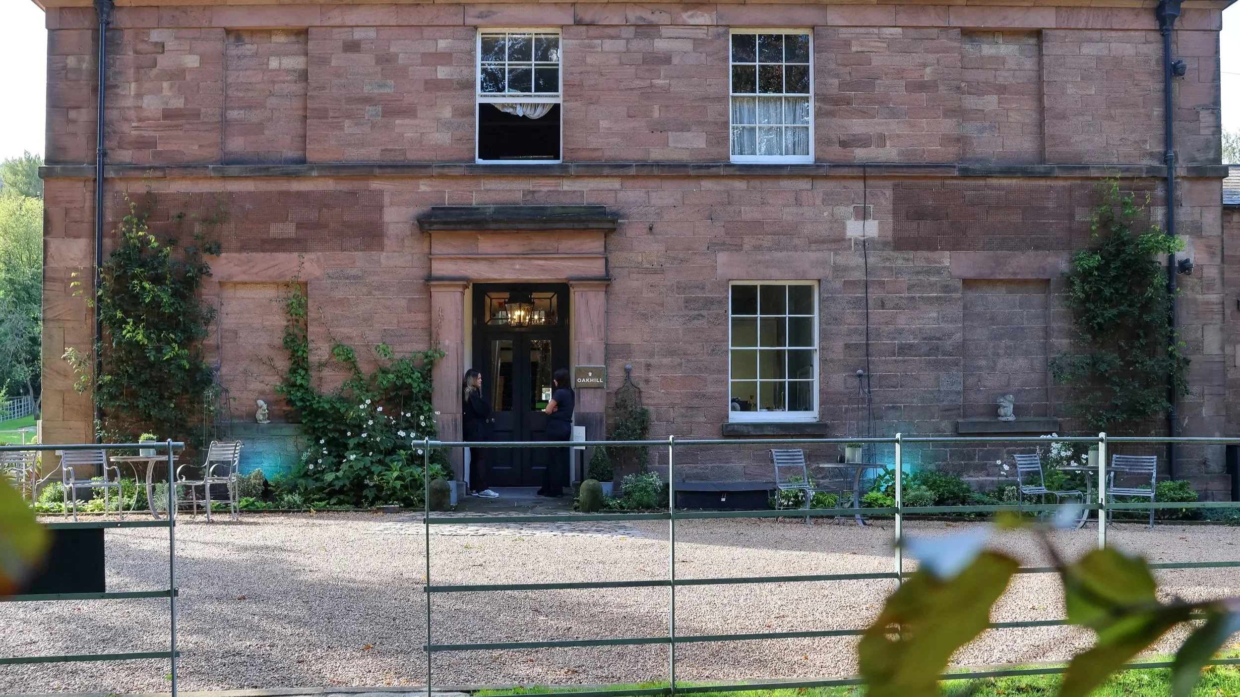 A brick building with several windows and a black door, with two women standing and speaking outside. There are plants and garden furniture in front of the building.