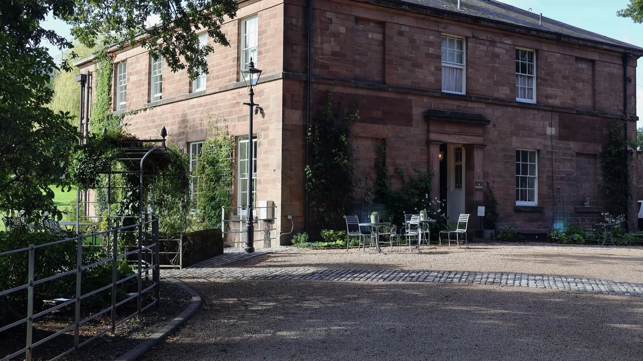A historic brick building with tall windows in a garden setting. There are outdoor chairs, a table, a black wrought-iron archway, and a lamppost in the scene, with greenery and trees surrounding the area.