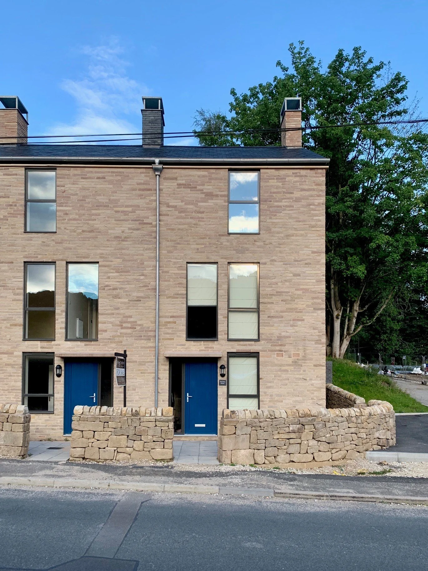 A modern multi-story brick building with blue doors, large windows, and a stone barrier in front. There are trees and a blue sky with some clouds in the background.