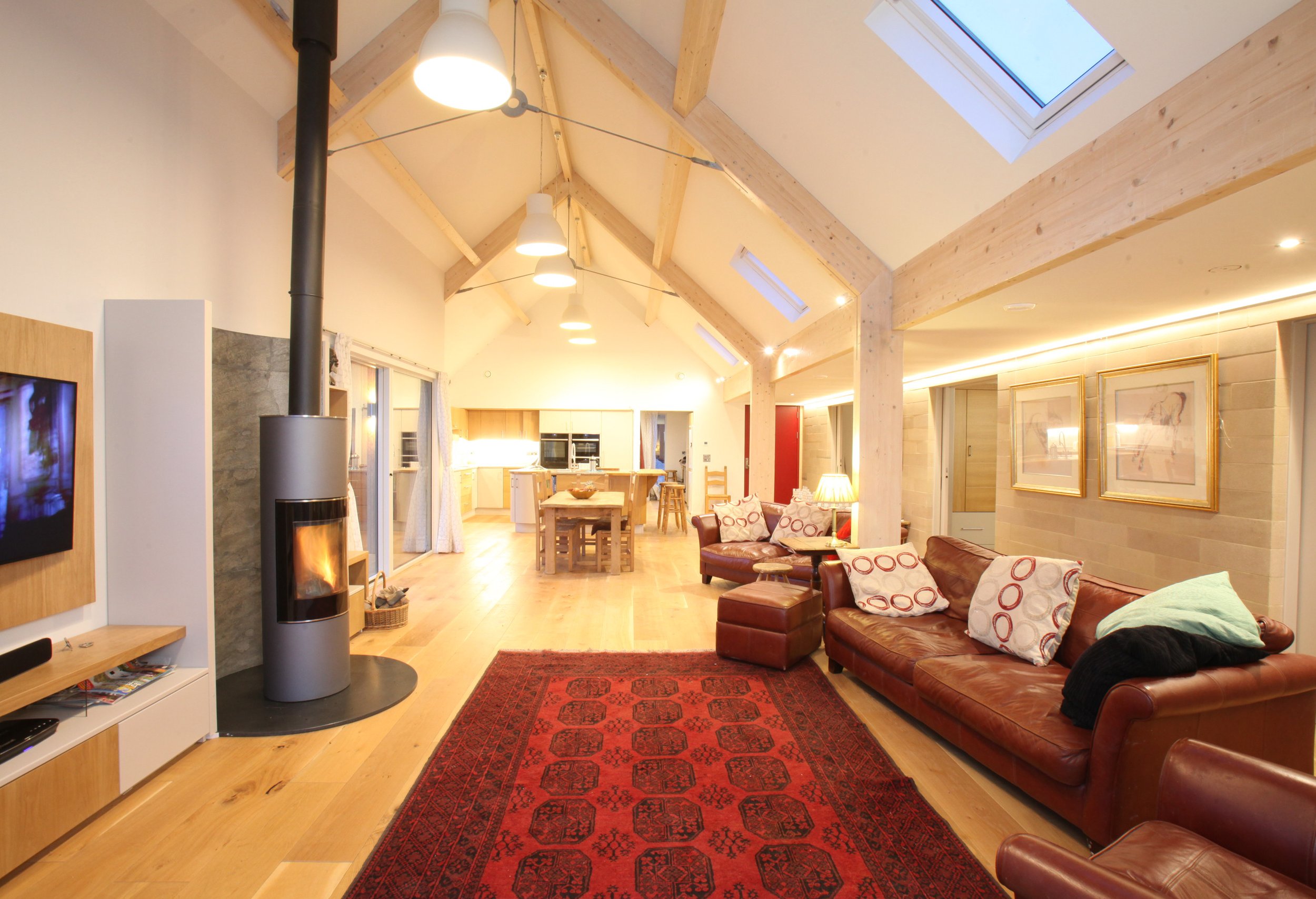 Living room with wooden beams, skylights, a red area rug, brown leather sofas with patterned pillows, a wooden dining table, and a fireplace, illuminated by ceiling lights and wall lighting.