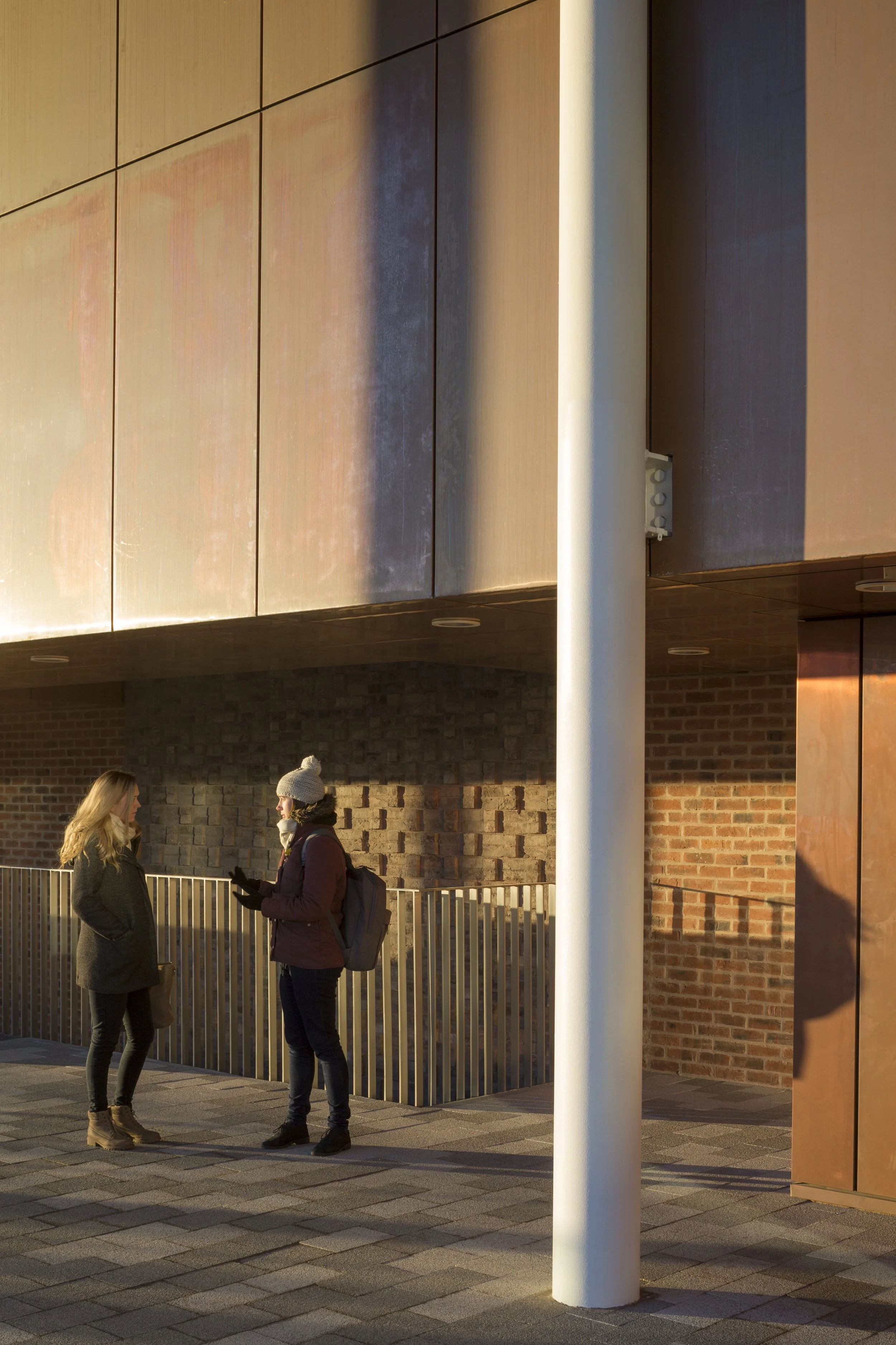 Two women standing and talking outdoors near a metal fence, with a brick wall and modern building facade in the background. One woman is wearing a dark coat and beige boots, the other is in a jacket and hat, holding a phone.