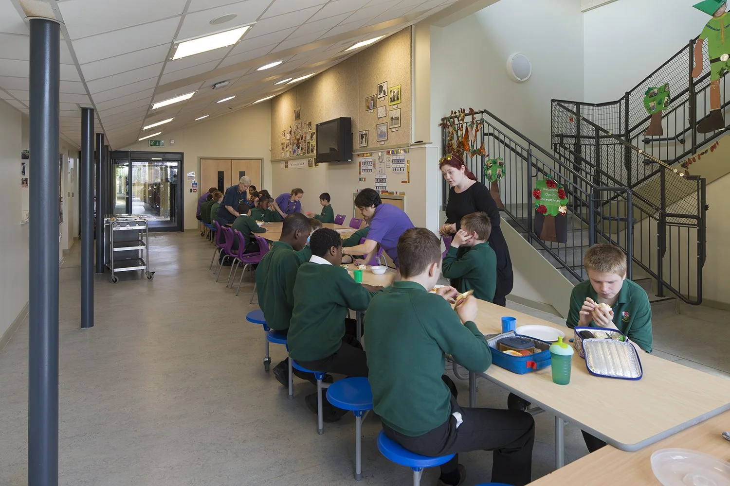 Students in green uniforms eating lunch in a school cafeteria decorated with Christmas crafts on the staircase railing.