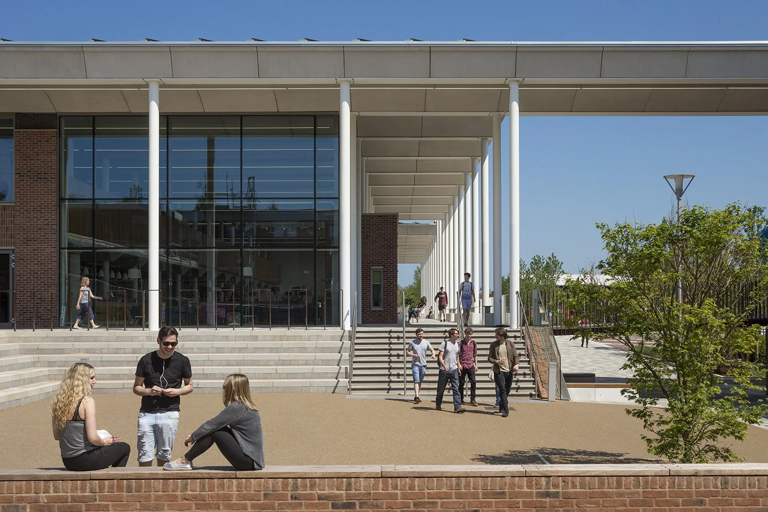 University campus with modern building featuring glass walls, white columns, and stairs, people walking and sitting outside on a sunny day.