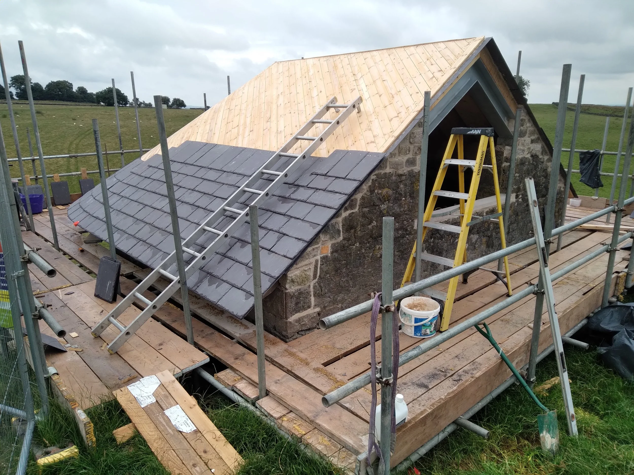 A small stone building with a partially constructed wooden roof, surrounded by scaffolding and construction tools in a rural field.