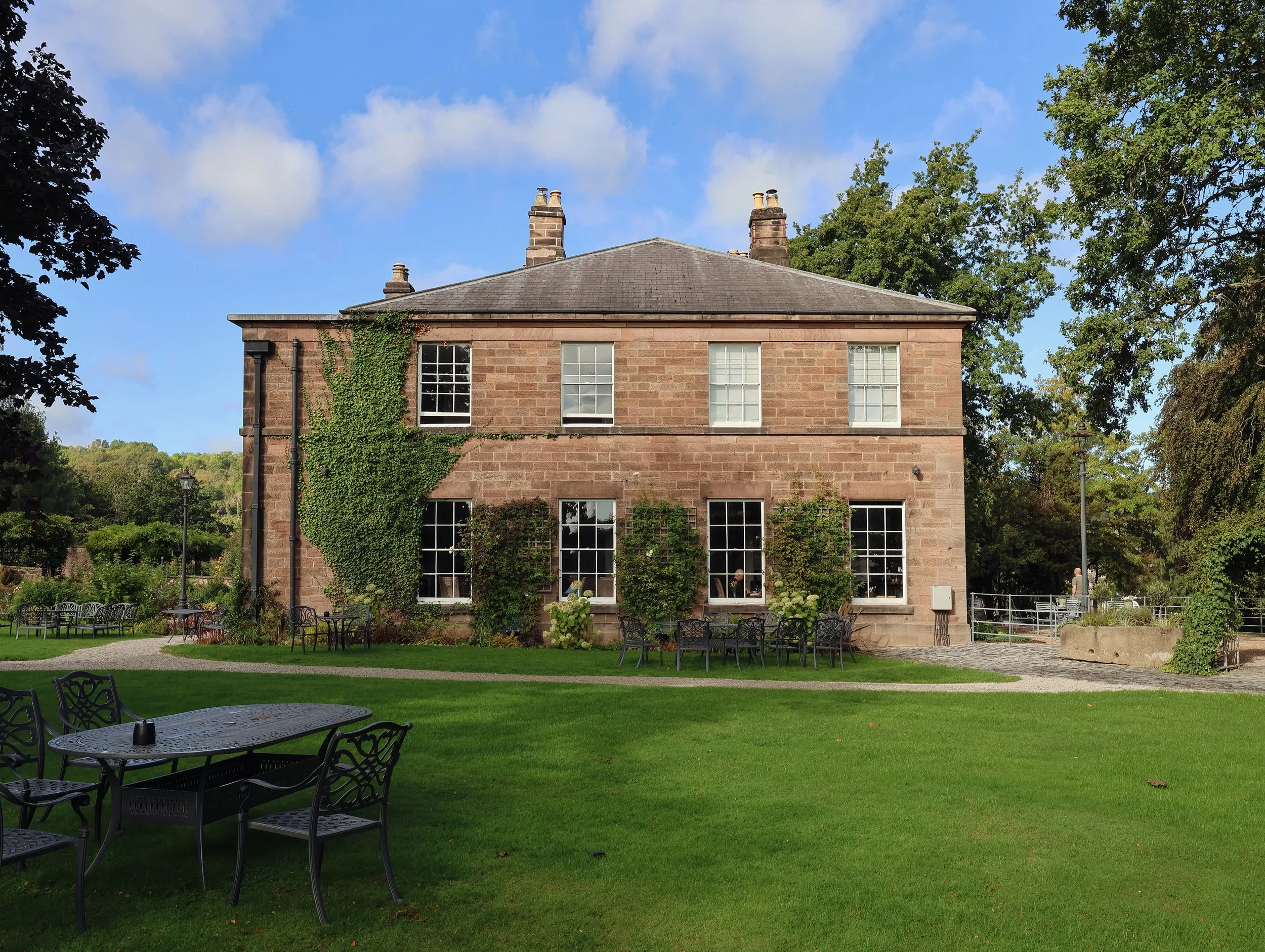A large red brick house with a sloped dark gray roof, multiple chimneys, and several large windows. The house has some green vines growing on one side. It is surrounded by a grassy lawn with outdoor tables and chairs, trees, and a clear blue sky with