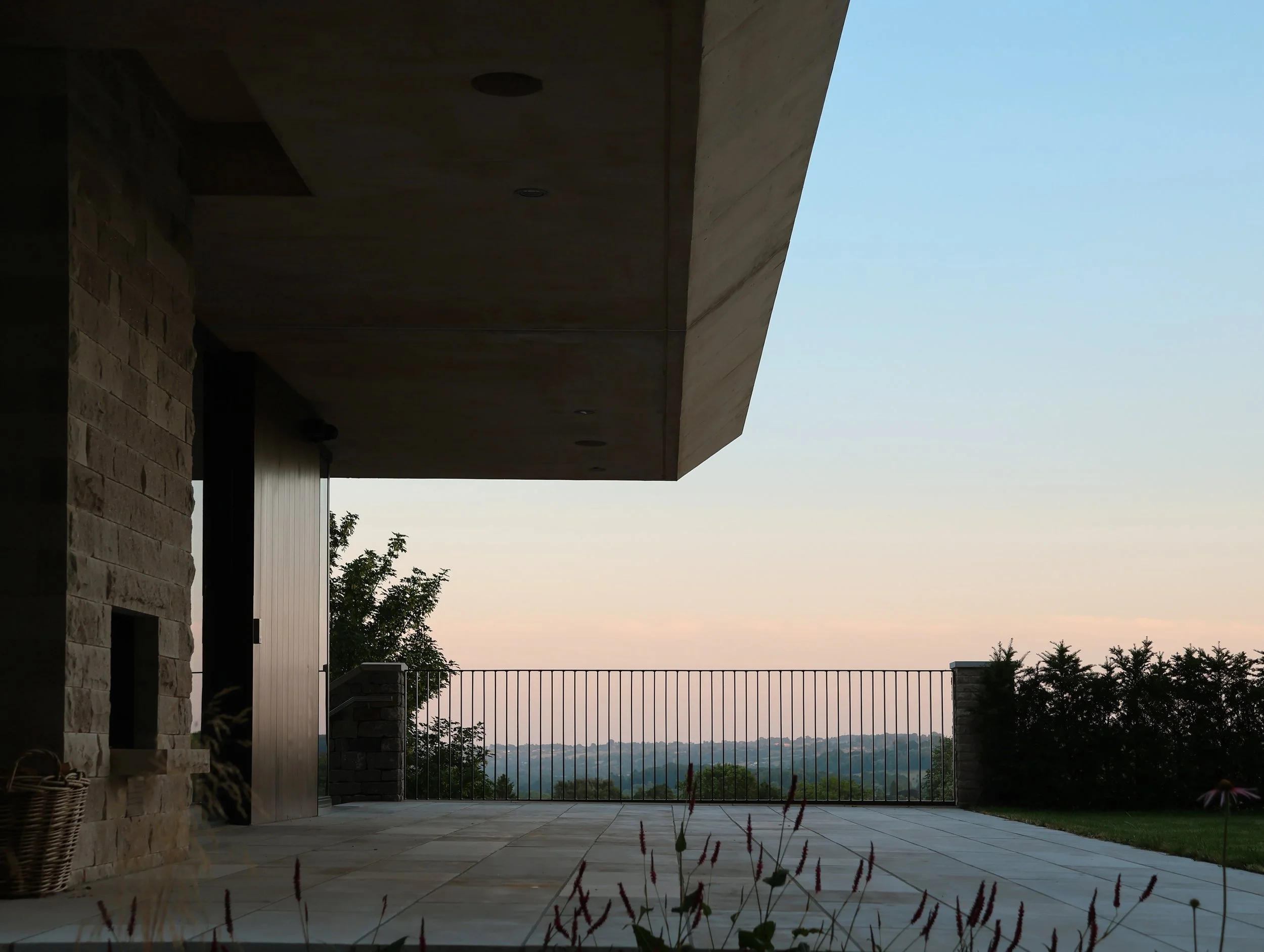 A patio with stone flooring and a metal railing, overlooking a landscape with trees and distant mountains at twilight.