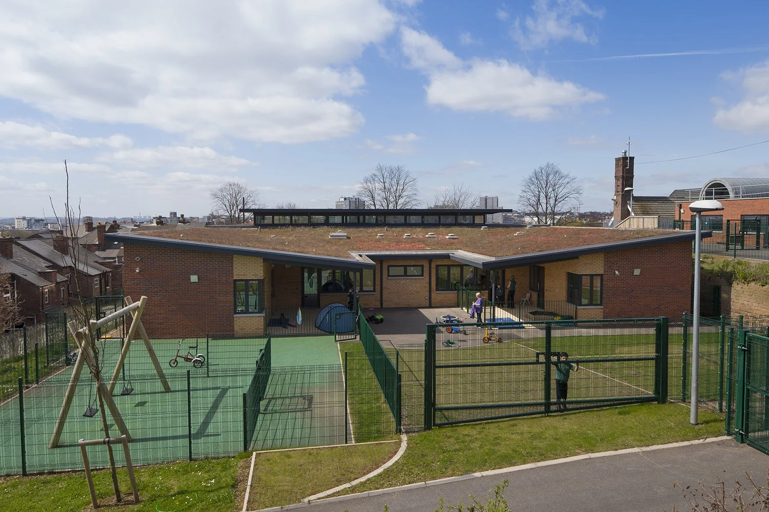 A modern, single-story brick building with a flat roof, situated behind a fenced playground area with children playing and adults nearby, under a partly cloudy sky.