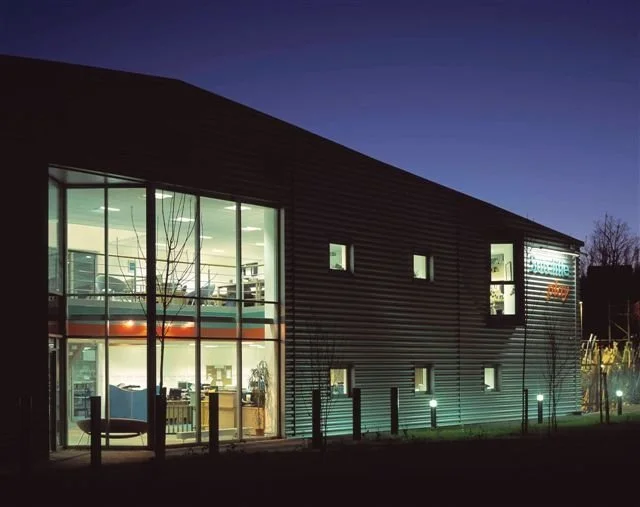 A modern building with large glass windows showing the interior lights and furniture inside, illuminated at night with exterior lighting along the sidewalk.