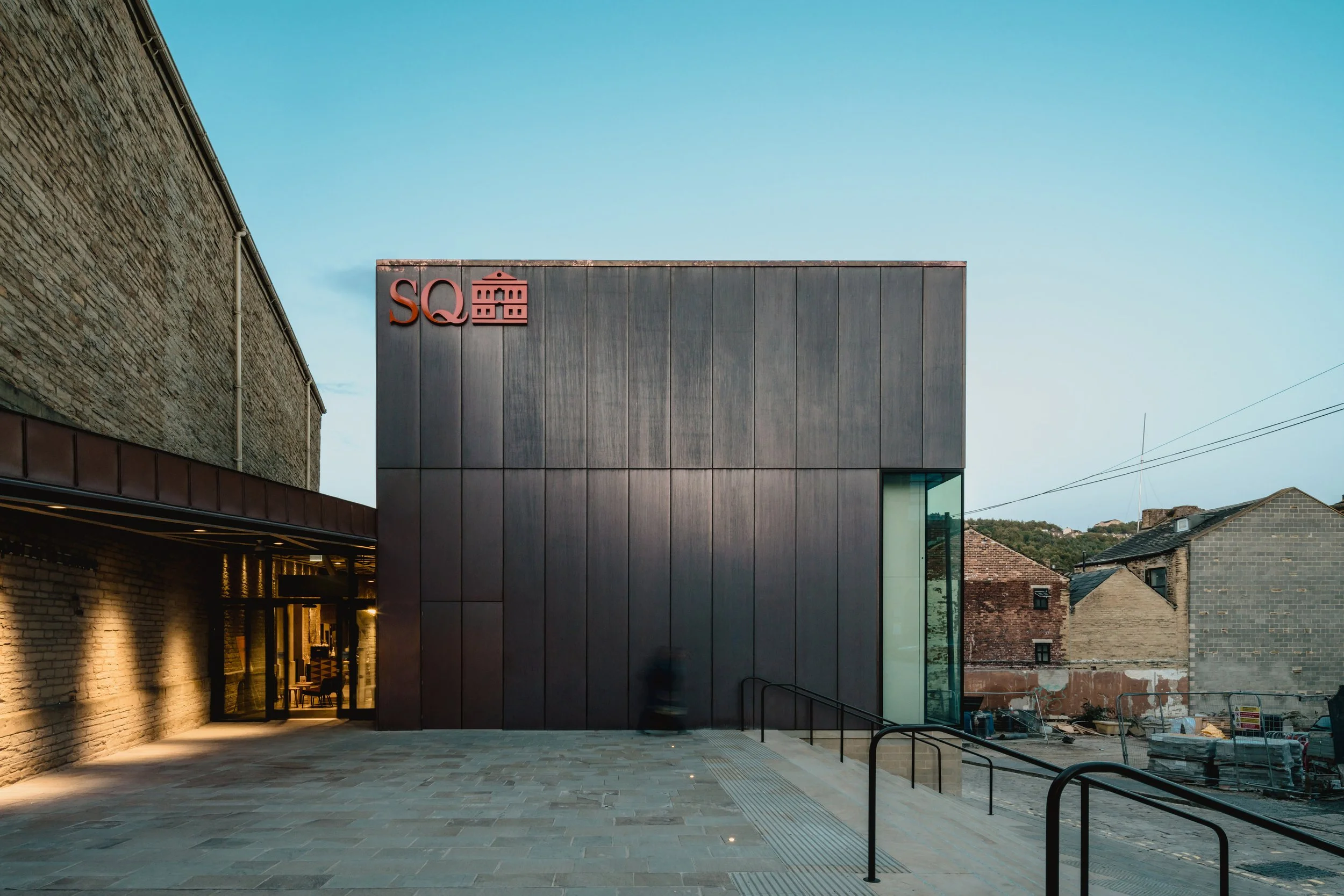 Modern building with a sign that reads 'SQ' and a stylized building icon, located next to an older brick building, under a clear sky.