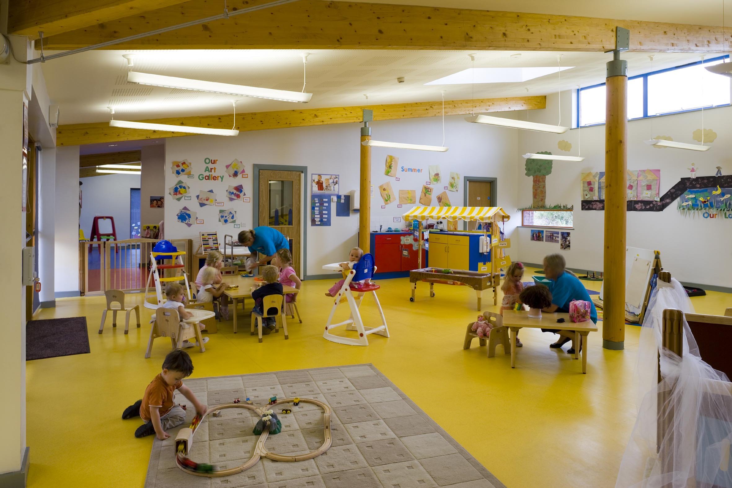 Children playing and drawing in a colorful preschool classroom with yellow flooring, wooden beams, and walls decorated with children's artwork.
