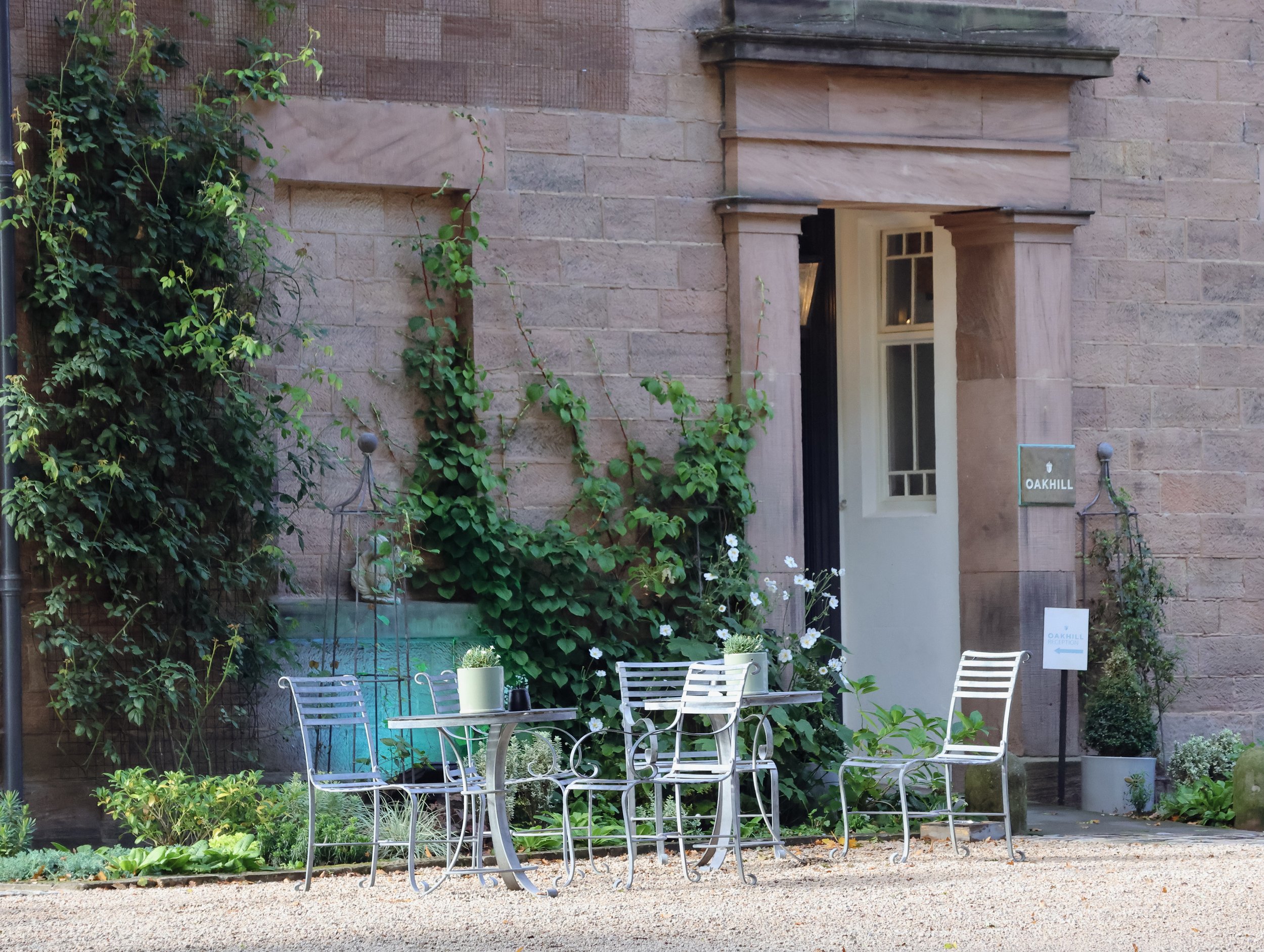 An outdoor patio area with metal tables and chairs, surrounded by greenery and plants, with a brick building in the background and a sign that reads 'OAKHILL'.