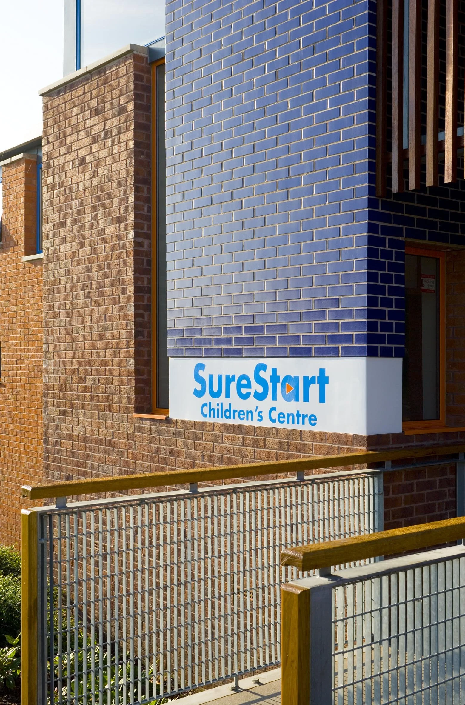 Exterior of a child care center with brick walls, a white sign displaying blue text 'SureStart Children's Centre', and a metal and wooden railing in the foreground.