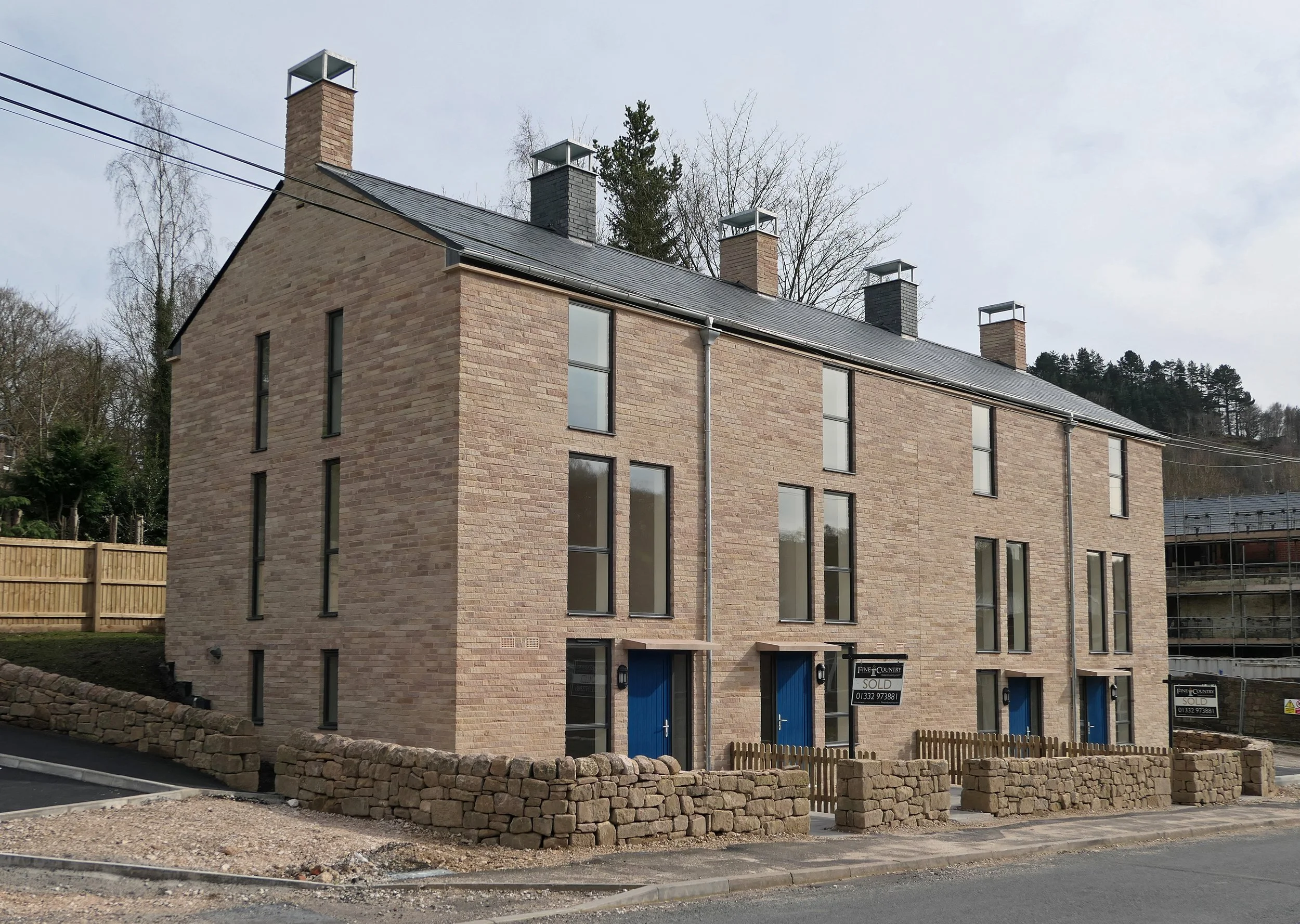 A modern multi-story brick building with large windows, blue doors, and multiple chimney stacks on the roof, surrounded by a stone wall and a small wooden fence, with trees and a cloudy sky in the background.