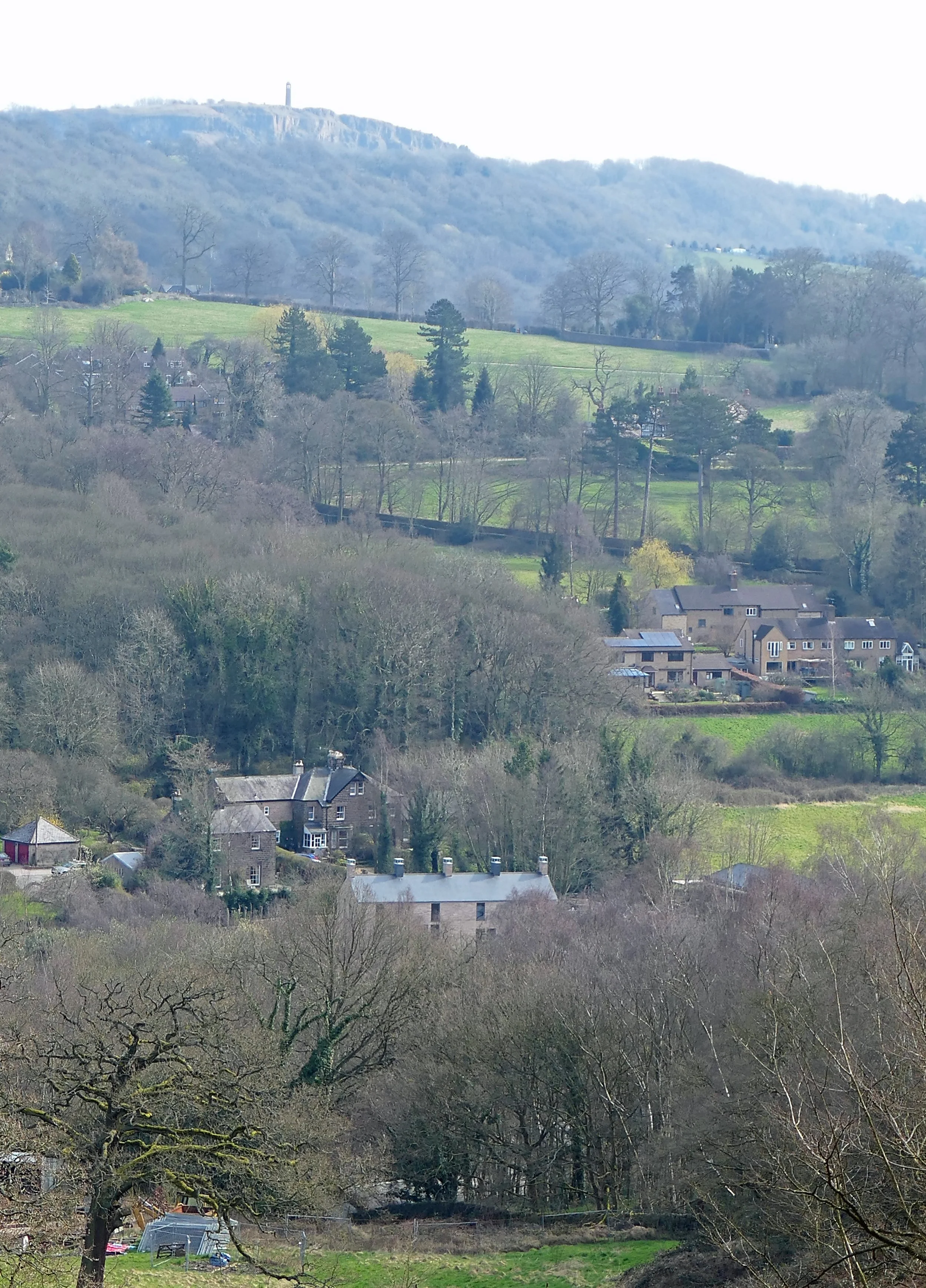 Hilly landscape with a mix of trees, houses, and greenery, and a mountain with a tower on top in the background.