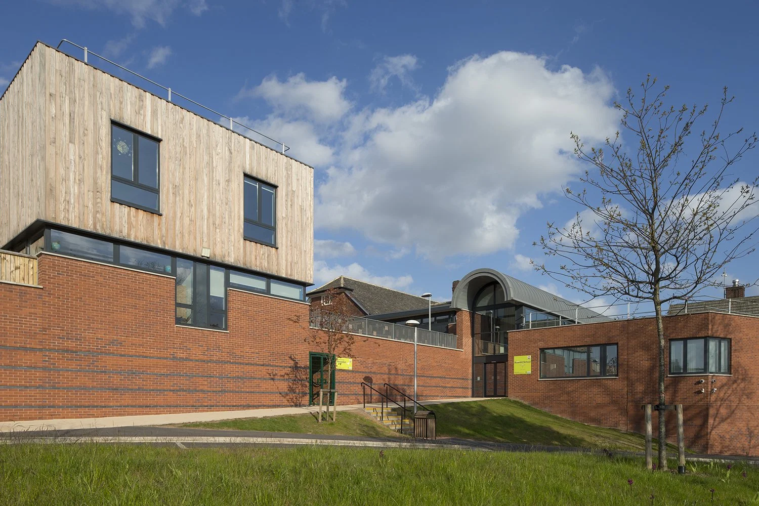 Modern building with brick and wood exterior, large windows, and a curved glass roof, set against a blue sky with clouds, with a small tree and grass in the foreground.