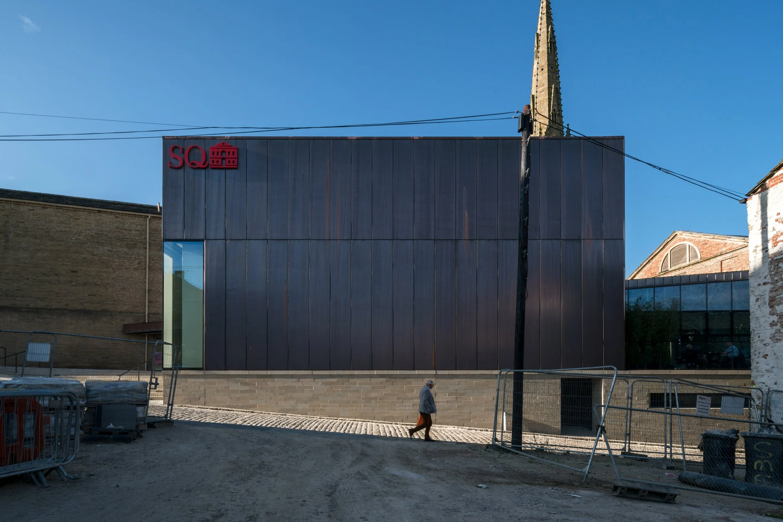 A modern building with a dark facade and large glass window, with a church steeple visible in the background under a clear blue sky. There are construction barriers and a person walking in the foreground.