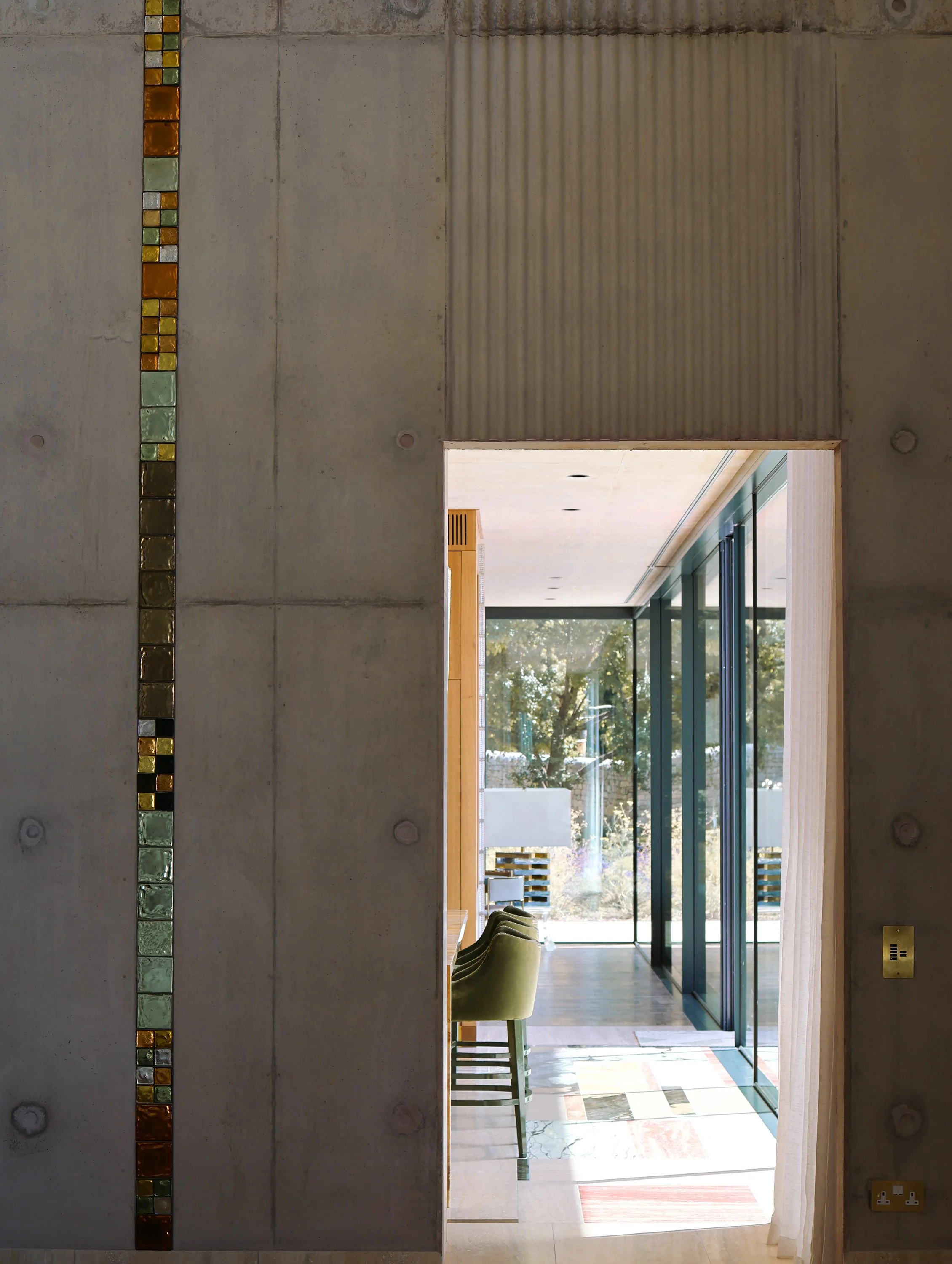 Interior view of a modern building, featuring a concrete wall with colorful glass block accents, an open doorway, and a hallway with a seating area and large windows showing trees outside.