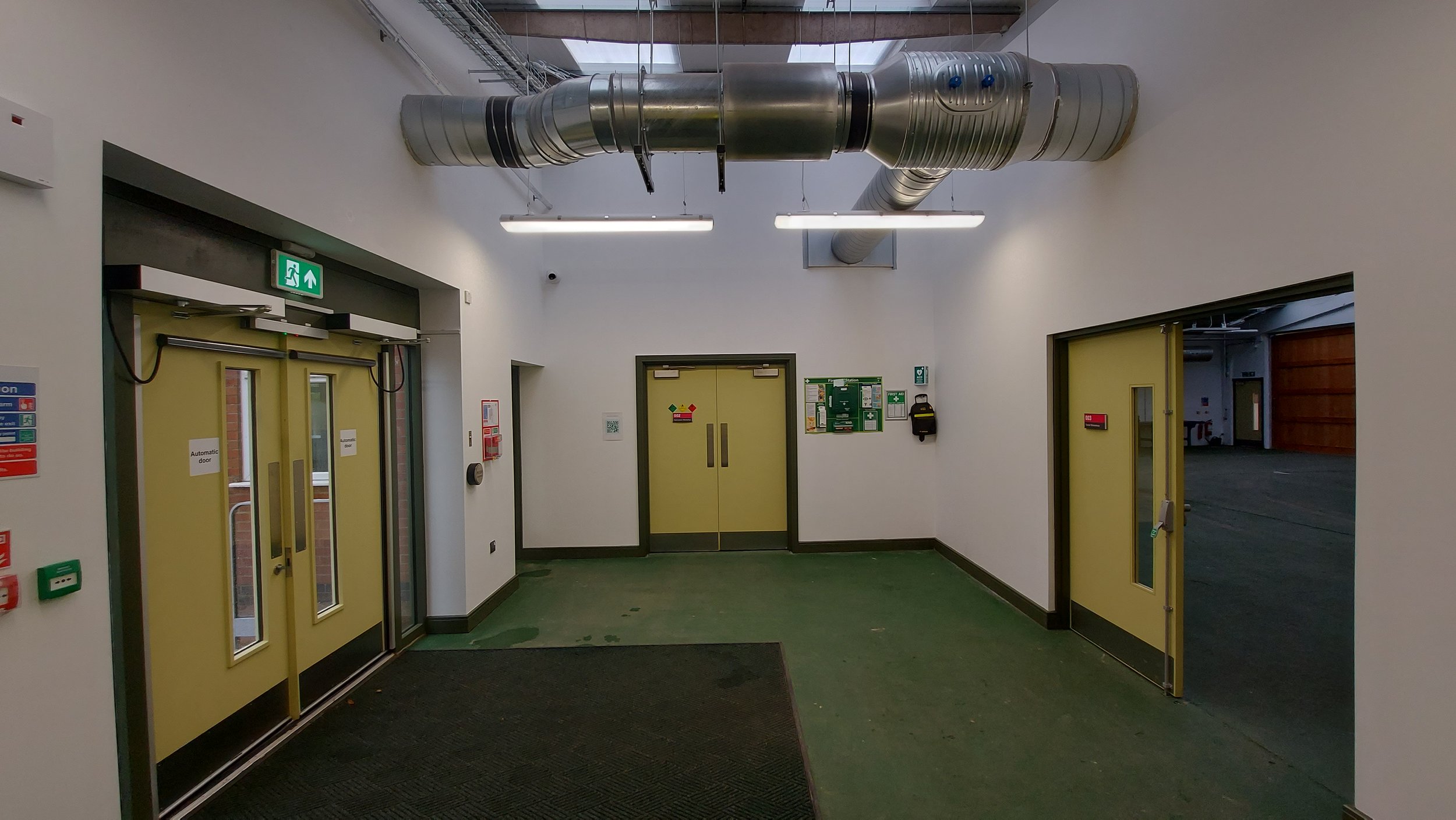 Interior of a building with yellow double doors, ventilation ductwork on the ceiling, emergency exit sign, and safety equipment.