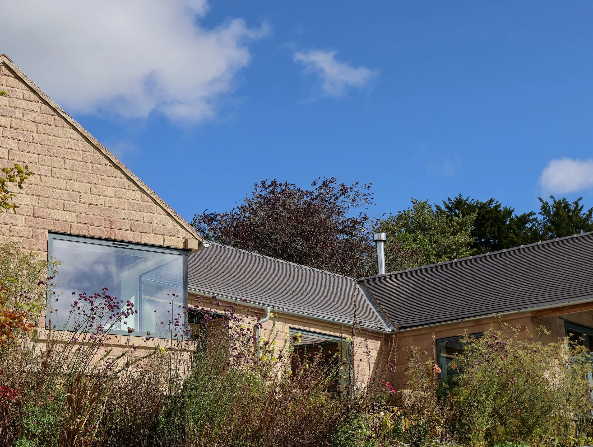 A residential house with a brick exterior, a large window, and a sloped roof with dark shingles. The garden in front has tall, flowering plants, and trees are visible in the background against a blue sky with some clouds.