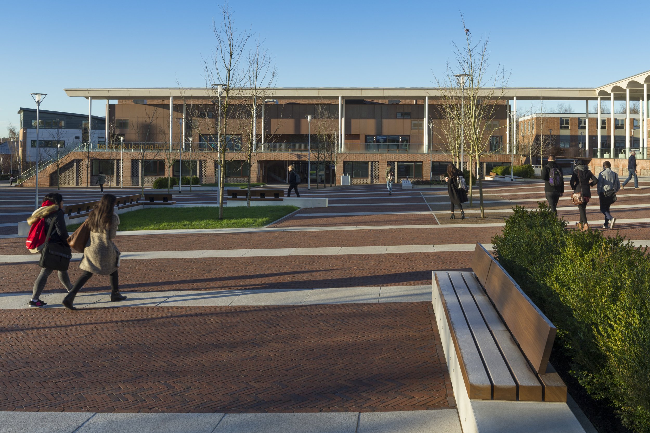 A modern campus building with a red brick facade, large windows, and a flat roof, surrounded by a plaza with benches, trees, and several people walking around on a sunny day.
