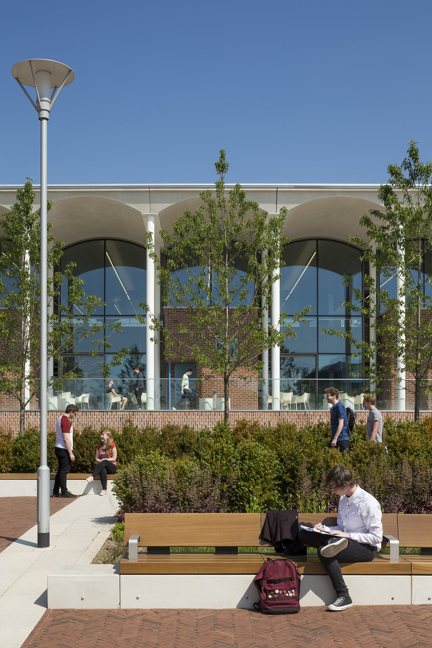 Students and pedestrians in an urban courtyard with landscaping, benches, and a modern building with large glass windows and arches.